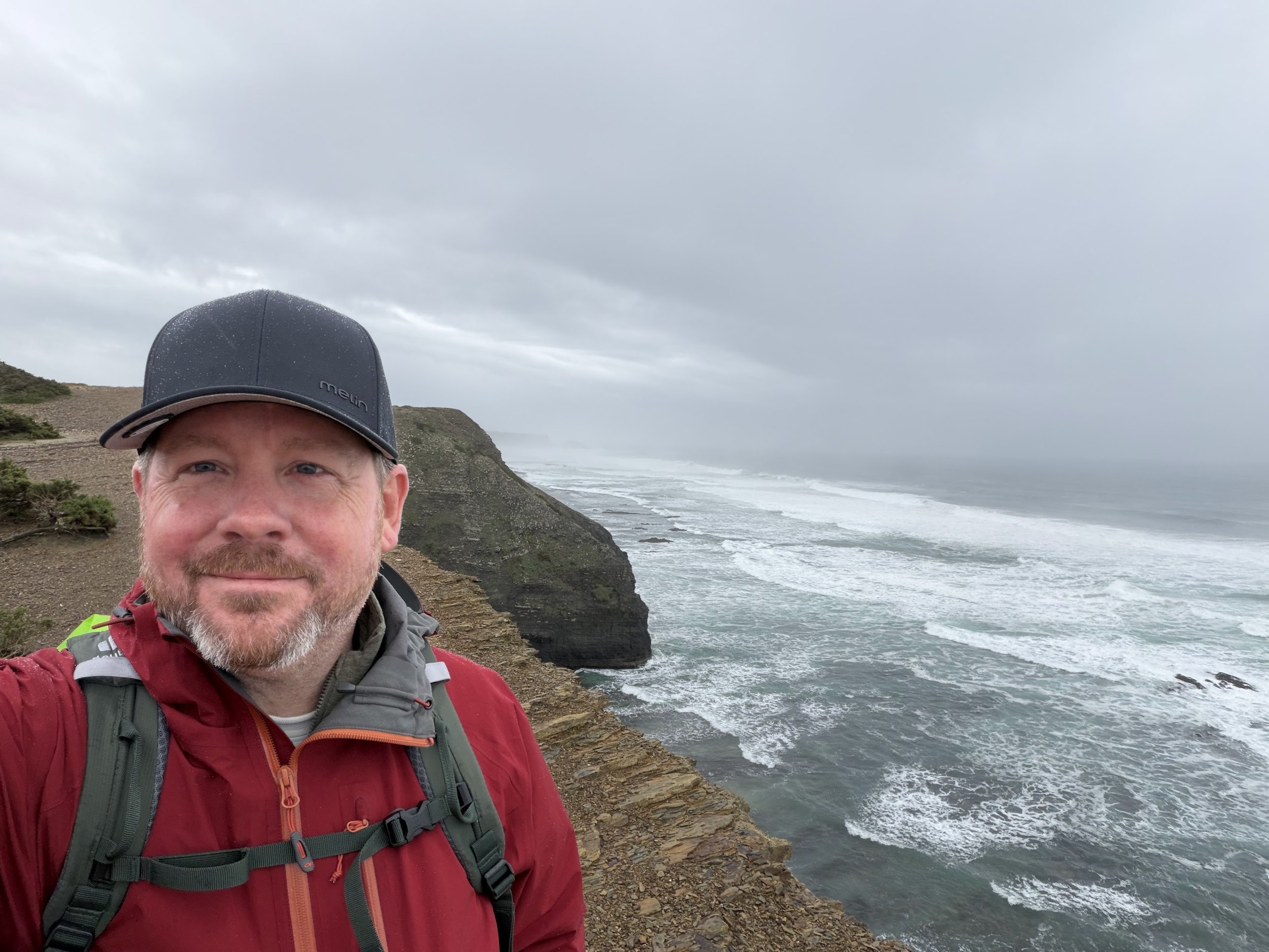 Brian, the founder of Formerge, taking a selfie in Portugal on a rocky coastal trail with cliffs and ocean waves in the background, under a cloudy sky.