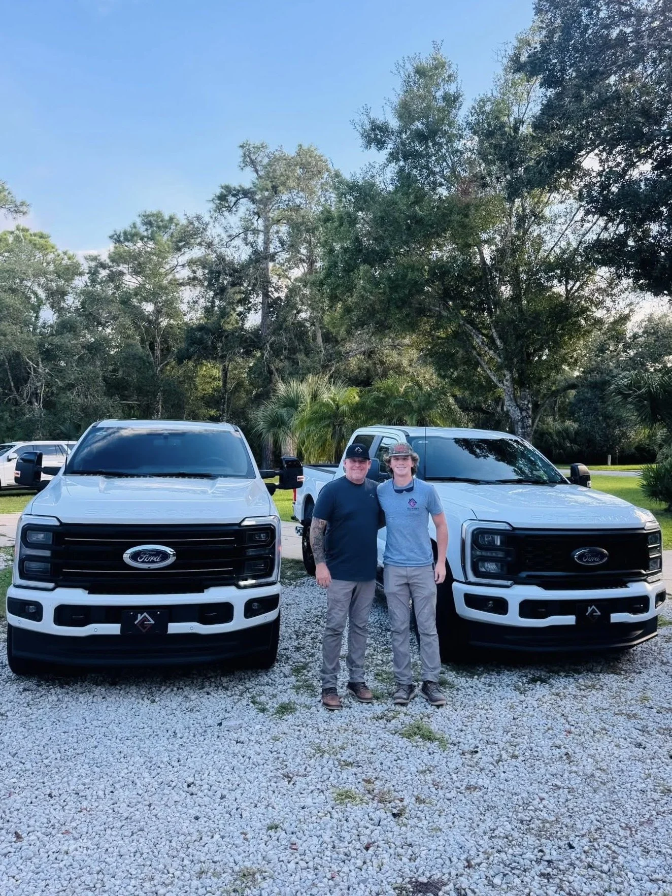 Two men standing in front of two white Ford trucks outdoors on a gravel area with trees and blue sky in the background.