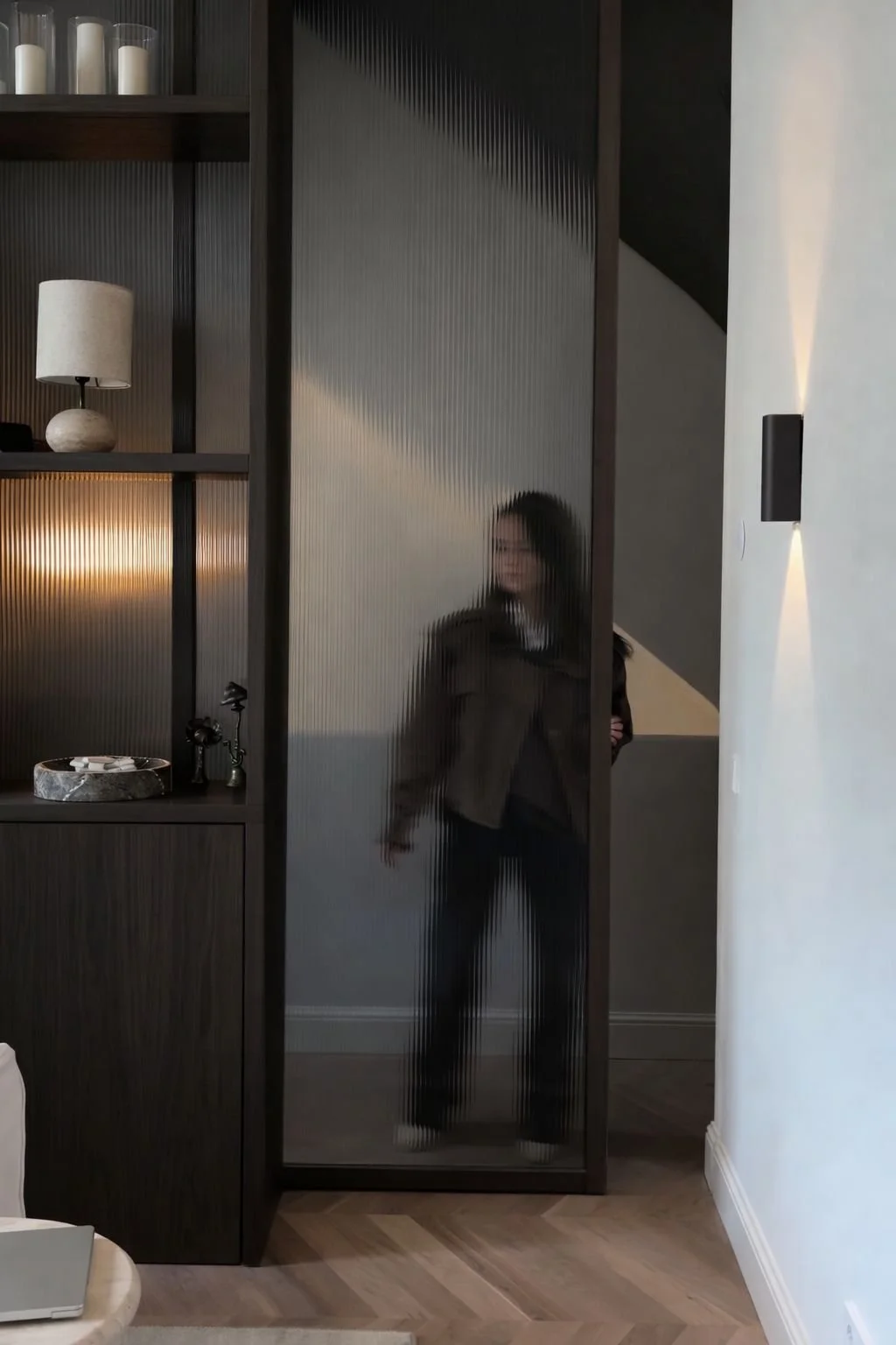 A woman with glasses and dark hair taking a selfie in a glass-structured door or partition inside a modern, minimalistic indoor space, with furniture and decorative objects visible behind her.