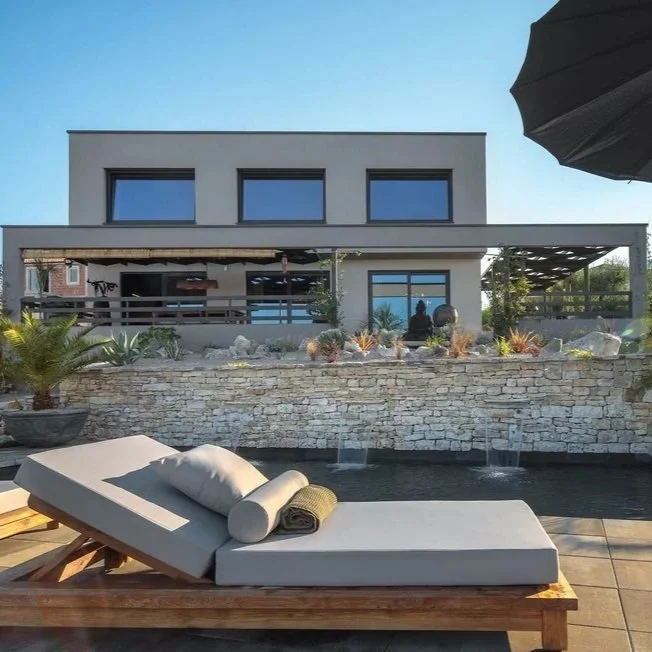 Modern house with poolside lounge chairs, surrounded by desert plants and trees, with mountain hills in the background during sunset.