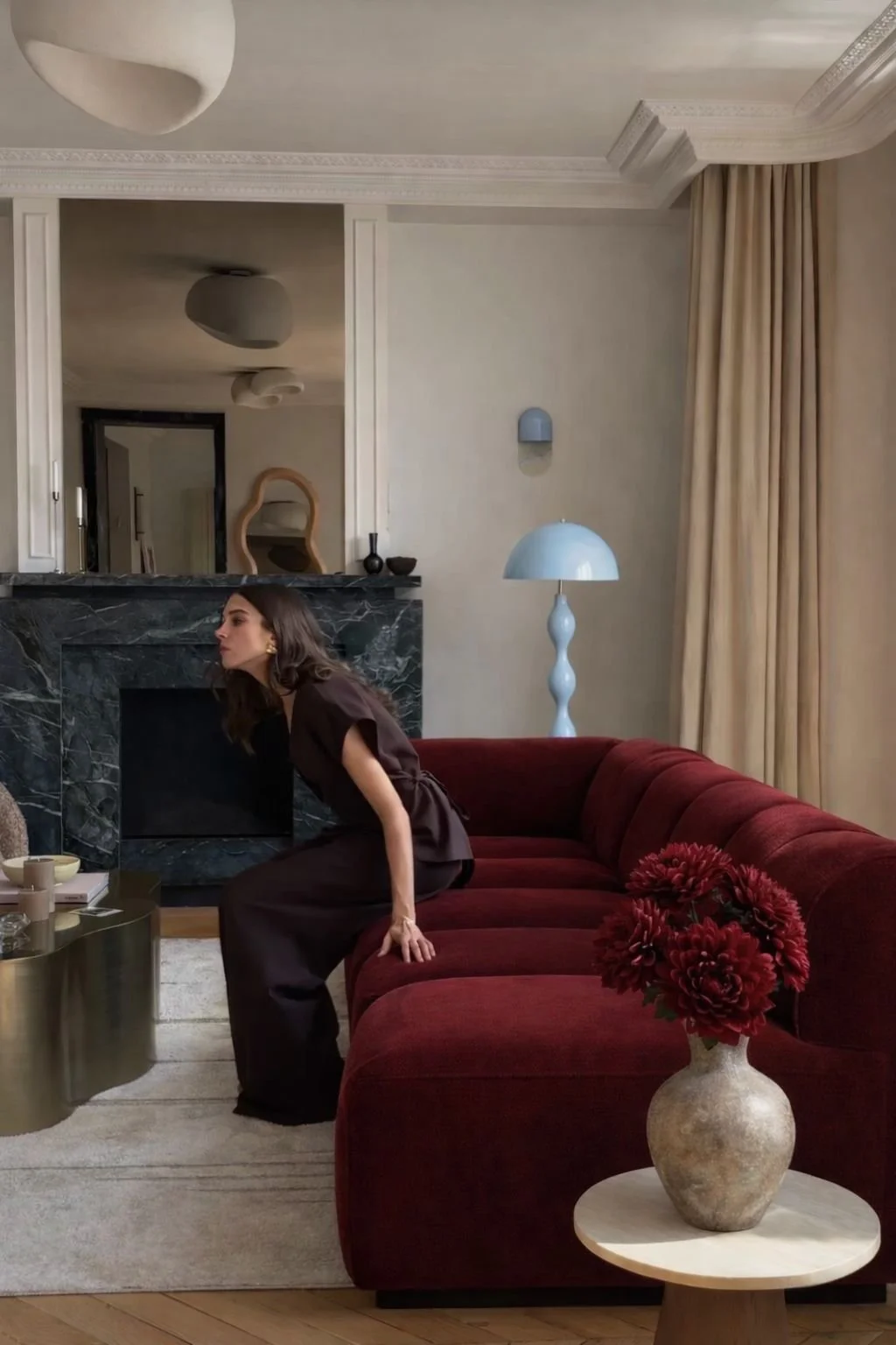 A woman with dark hair wearing a dark outfit sits on a burgundy sofa in a stylish living room with a modern interior design, a black marble fireplace, a marble coffee table, a white table lamp, and a vase of red flowers in the foreground.