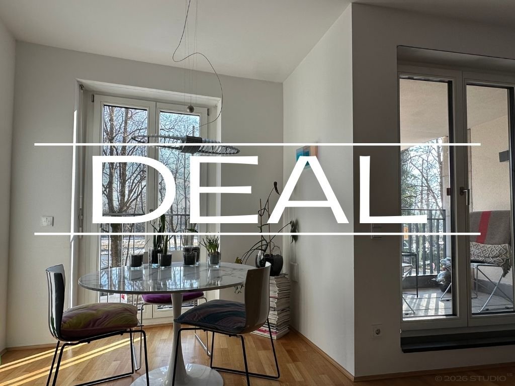 View of a bright dining area with a round glass table, four chairs, and glass doors leading to a balcony. There are potted plants on the table and a stack of magazines or papers beside the wall.