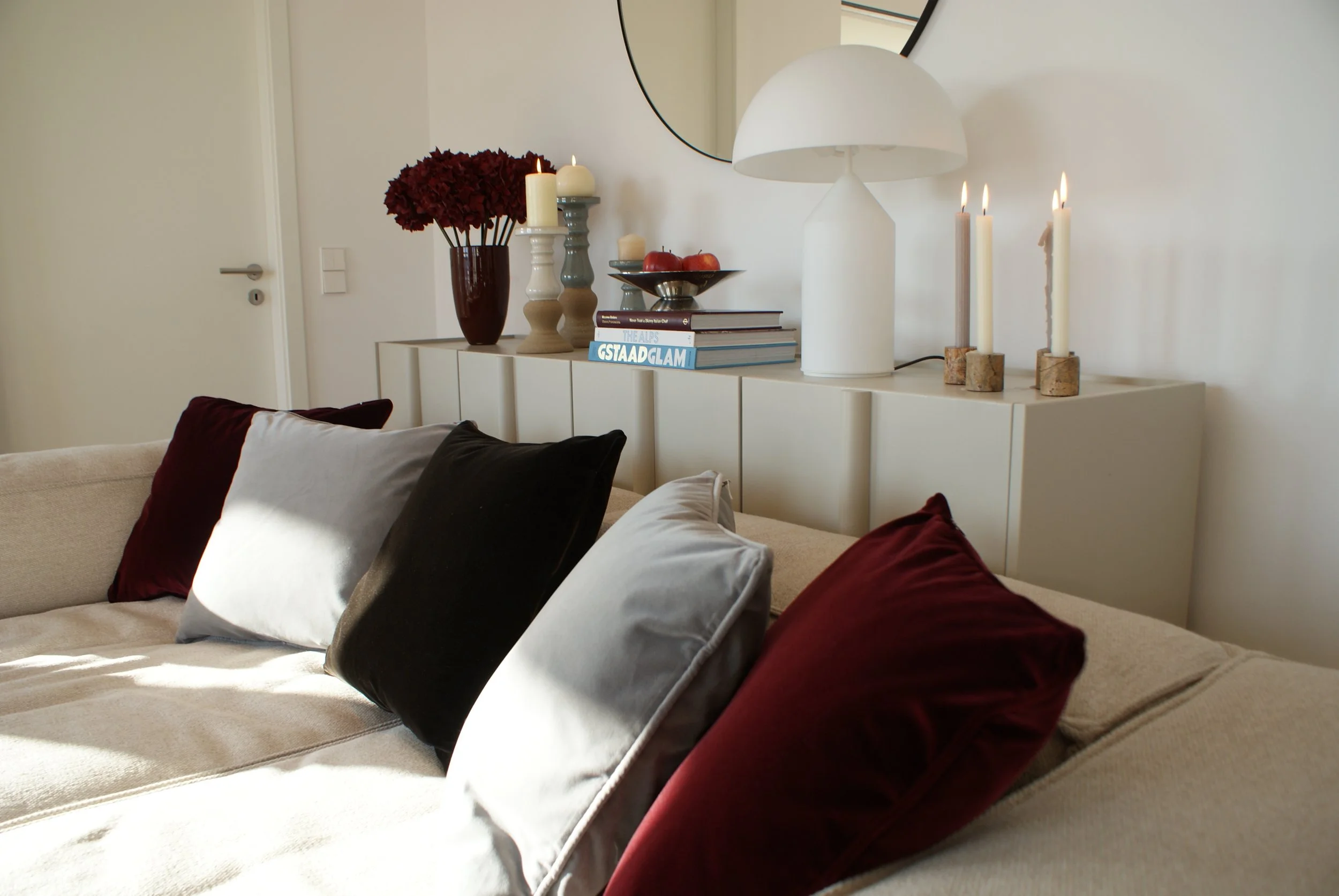 Living room with beige sofa adorned with red, black, gray, and white pillows. Behind the sofa, a white sideboard holds red flowers in a dark vase, candles, books, a tray with apples, and a large white lamp. A round mirror is mounted on the wall above the sideboard. Candles are lit, creating a cozy ambiance.