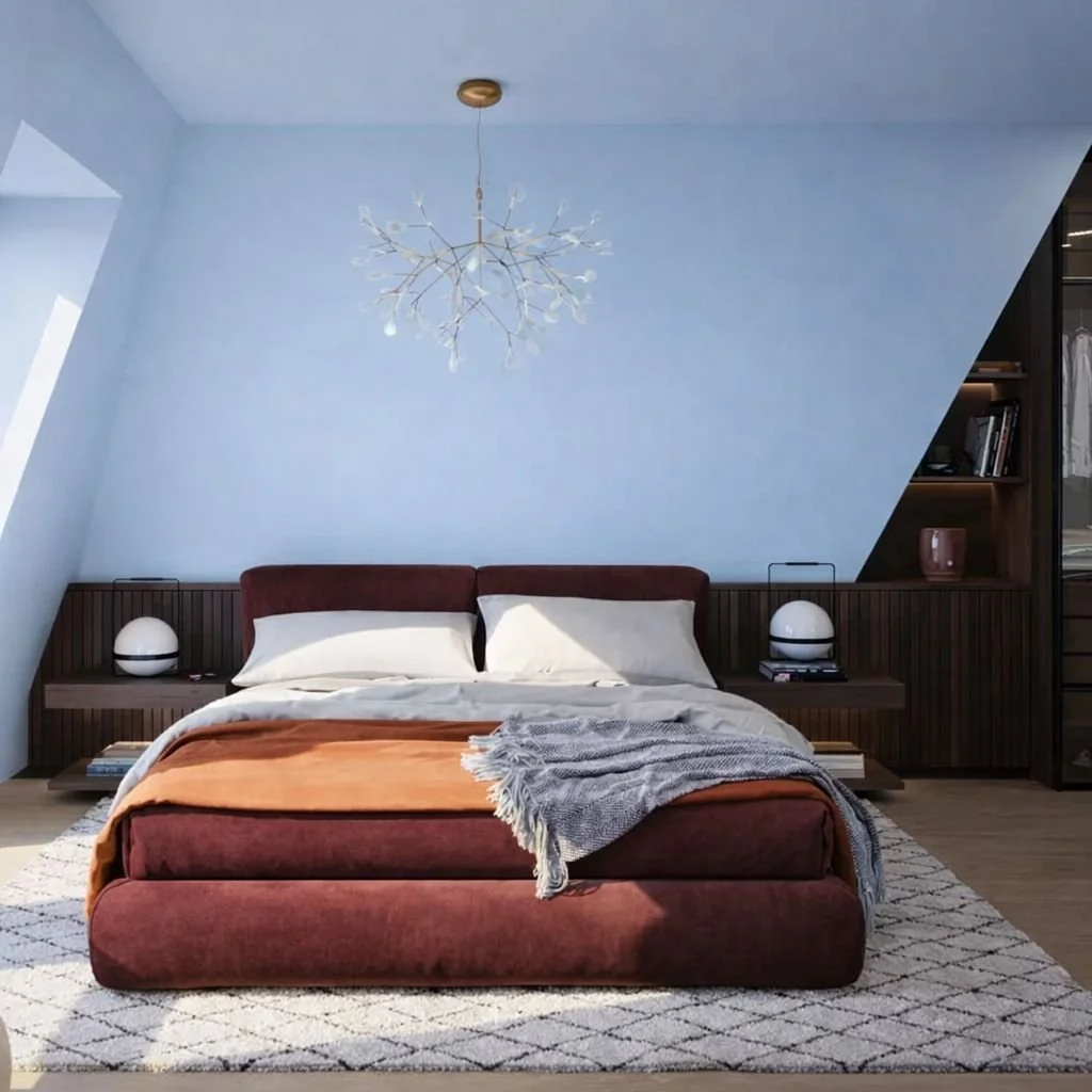 Modern bedroom with a burgundy bed, white pillows, beige and rust-colored blankets, a rug, and built-in wooden shelves on the right side. A chandelier hangs from the ceiling, and sunlight enters through a window on the left.