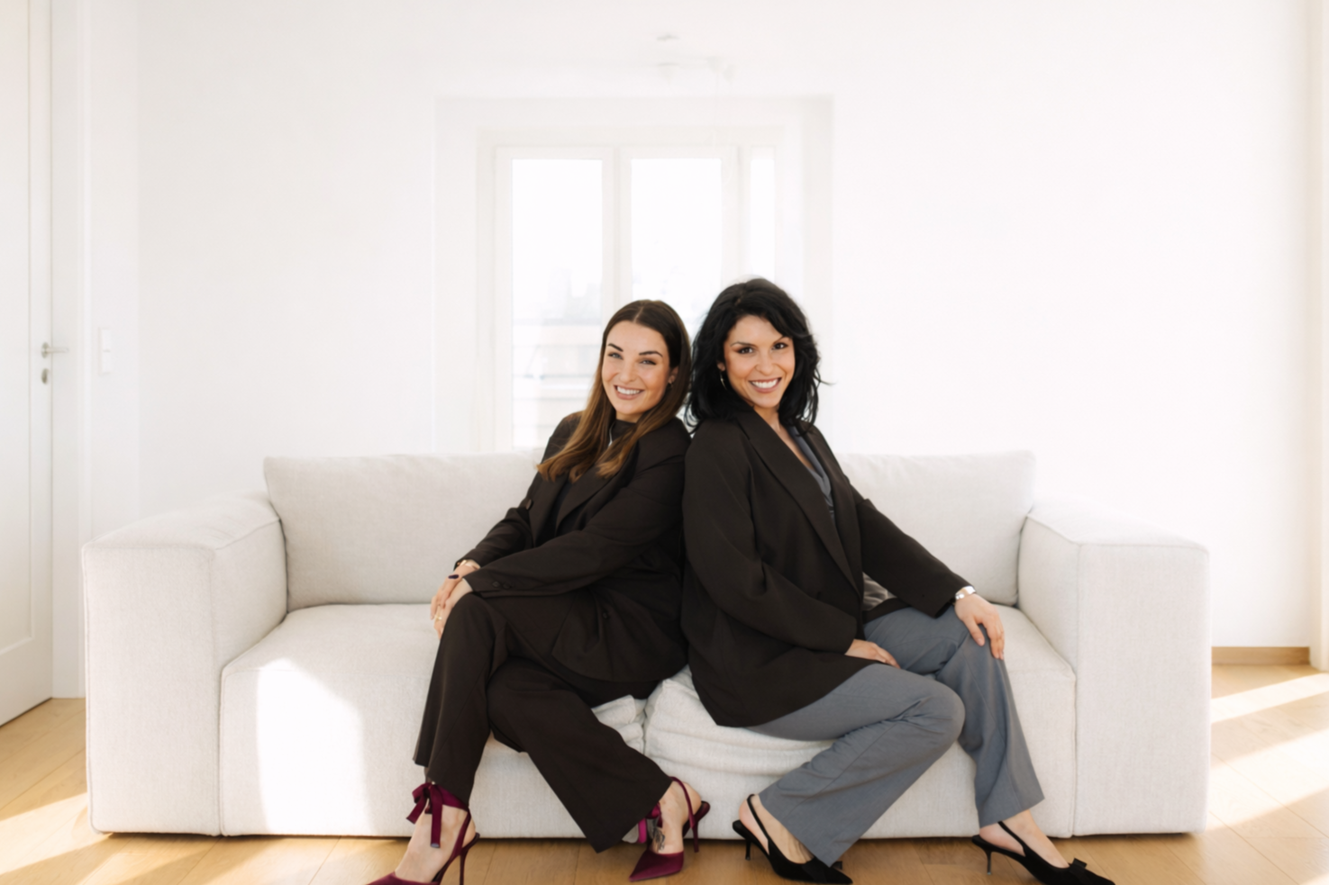 Two women in business attire sitting back-to-back on a light-colored sofa in a bright, minimalist room, smiling at the camera.