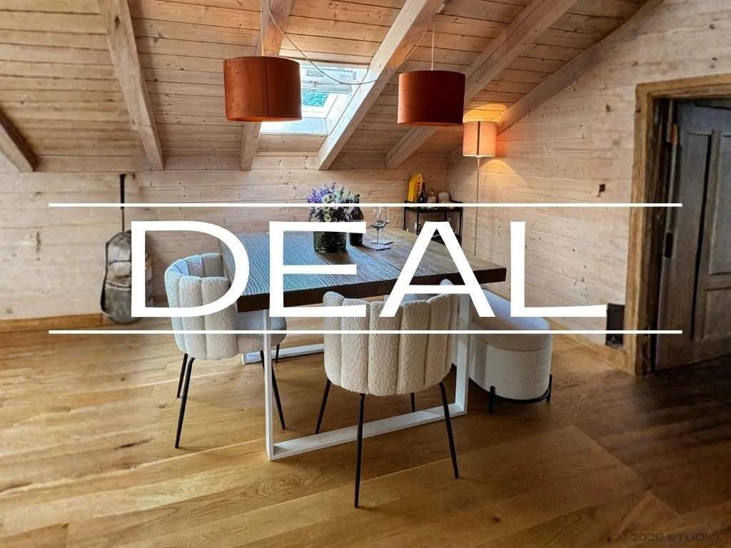 A cozy dining area in a mountain cabin with wooden walls and ceiling, featuring a large wooden table, six upholstered chairs, hanging pendant lights, and a small side table with a lamp, with a skylight in the ceiling bringing in natural light.