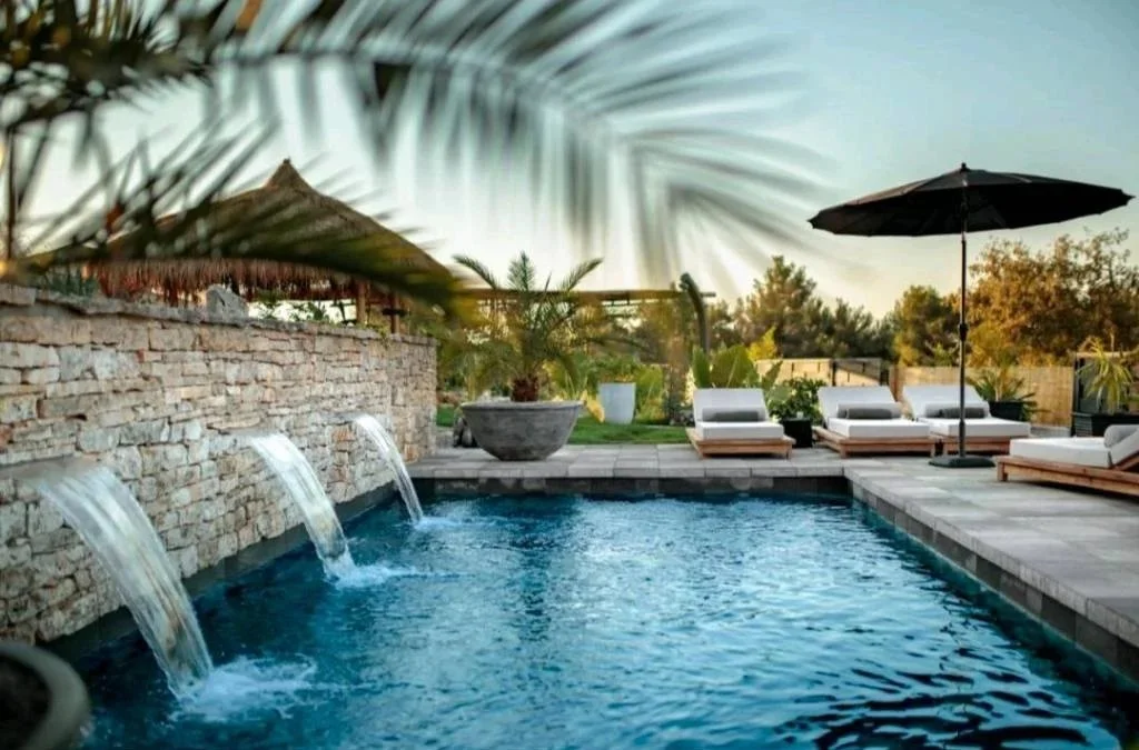 Modern house with poolside lounge chairs, surrounded by desert plants and trees, with mountain hills in the background during sunset.