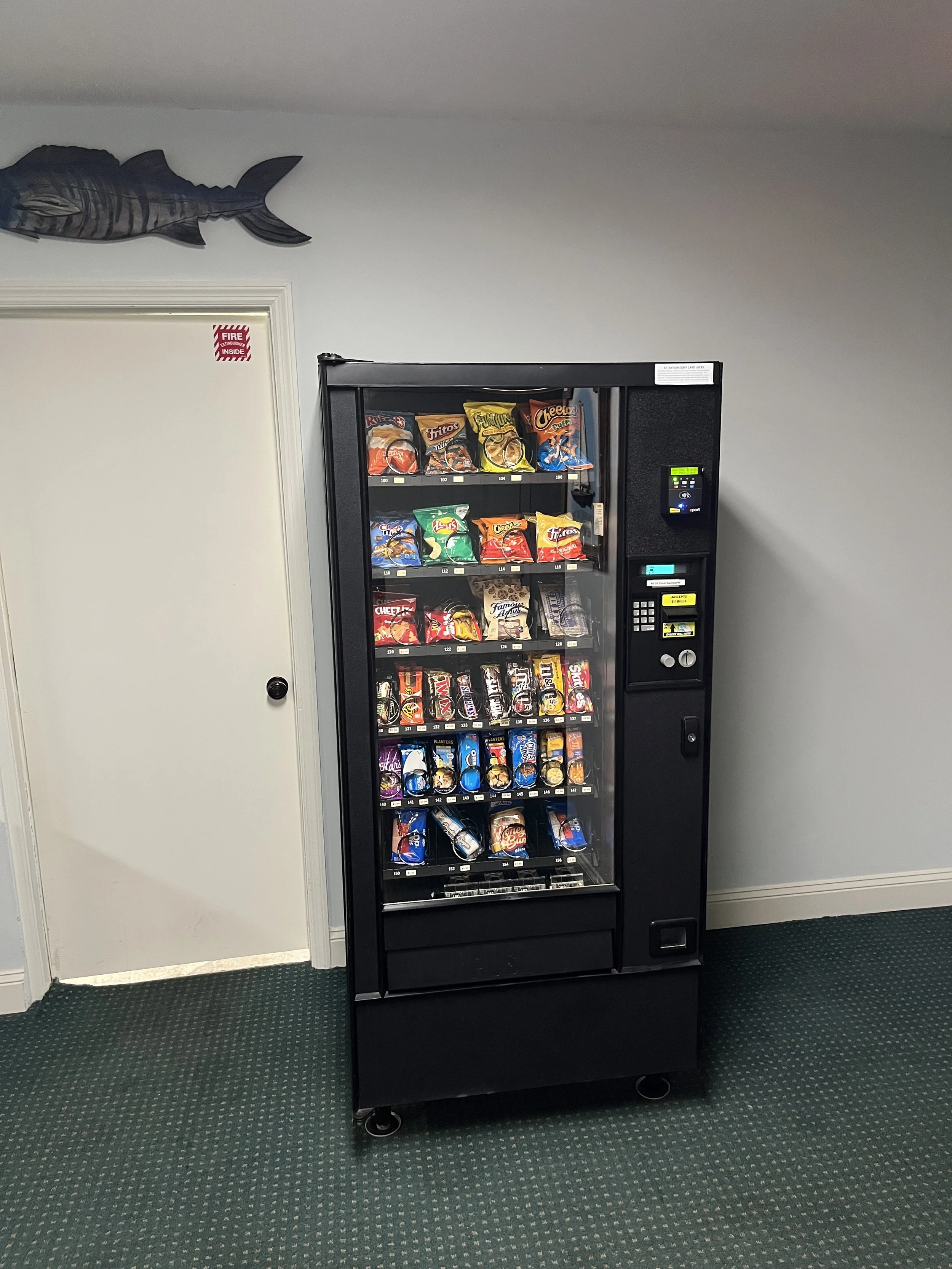 A vending machine filled with snacks, positioned next to a white door with a small red fire safety sticker. A fish-shaped decoration is mounted on the wall above the door.