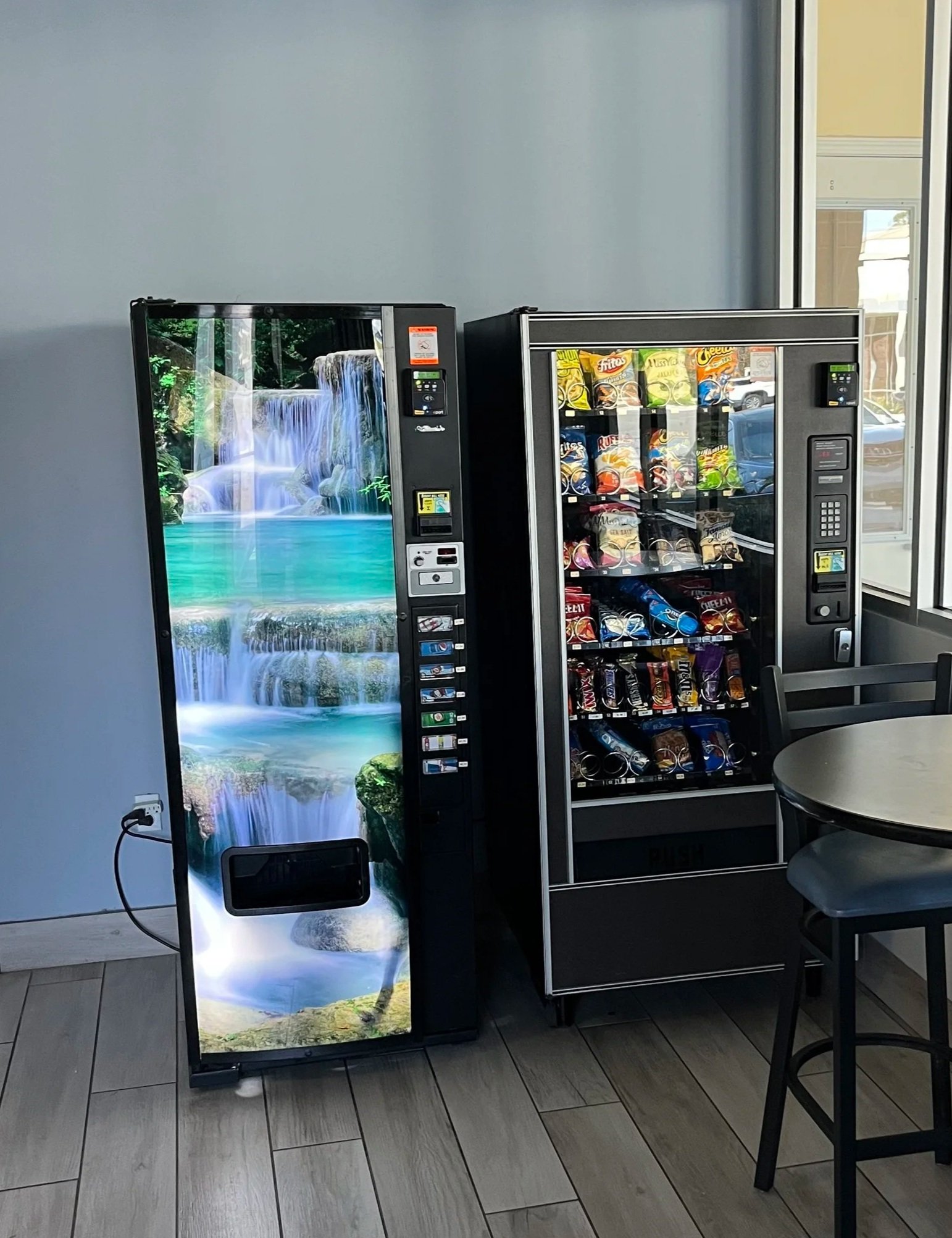 A vending machine with a nature scene of a waterfall and river on its door, next to another vending machine stocked with snacks, in a room with a round table and chairs.