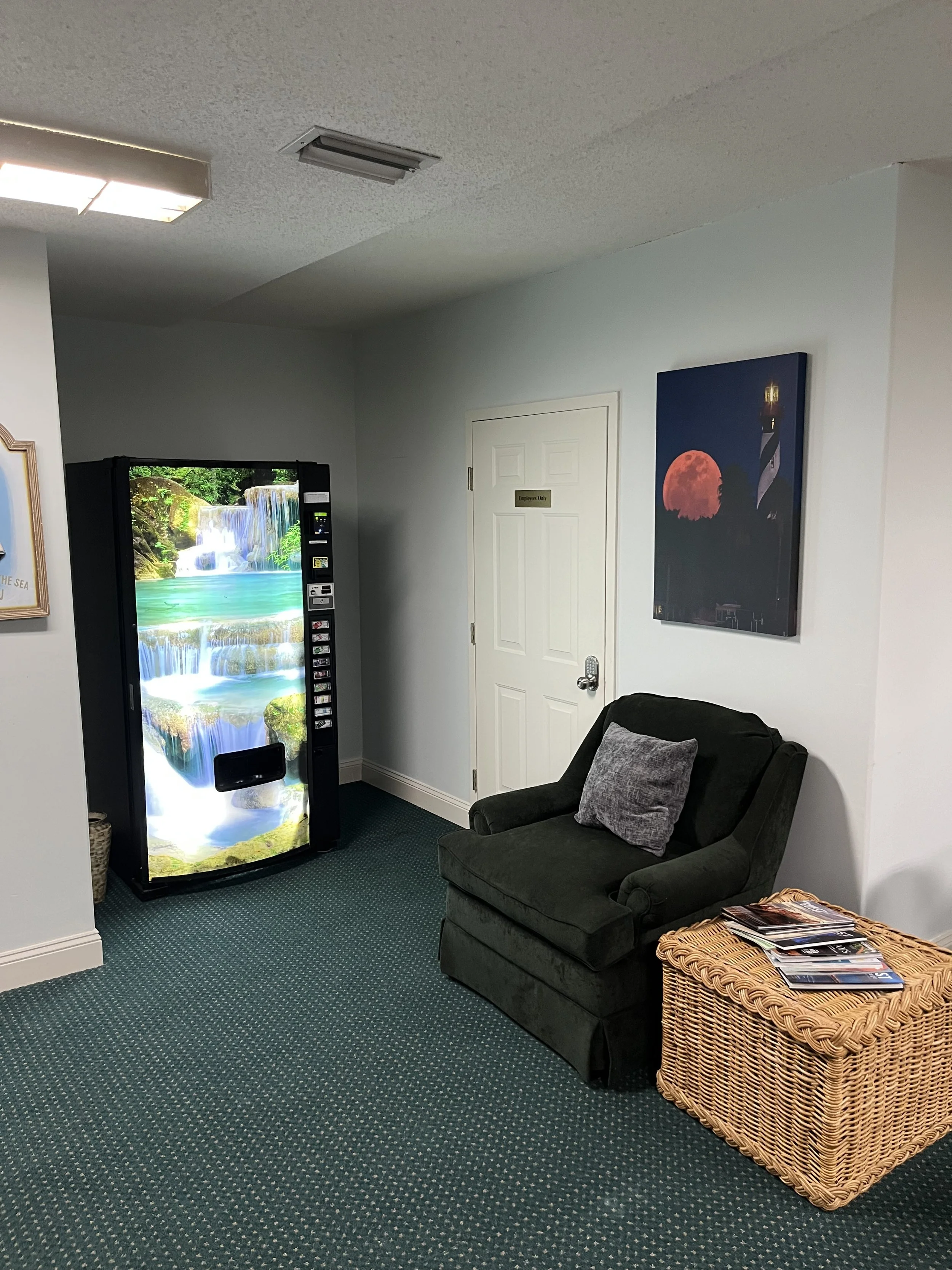 A waiting room with a dark green armchair, gray pillow, wicker table with magazines, vending machine with a nature scene, white door labeled "Employees Only," and wall art with a lighthouse and a red moon.
