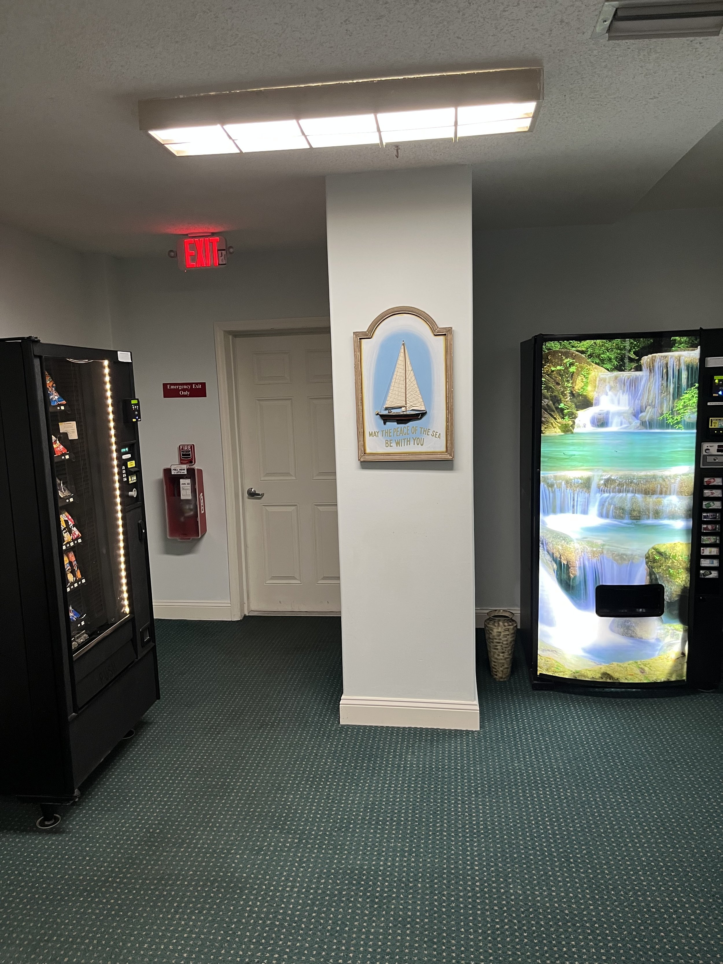 Vending machines in a room with a white door, a framed artwork of a sailboat with the text "May the peace of the sea be with you," a basket, and an exit sign overhead.