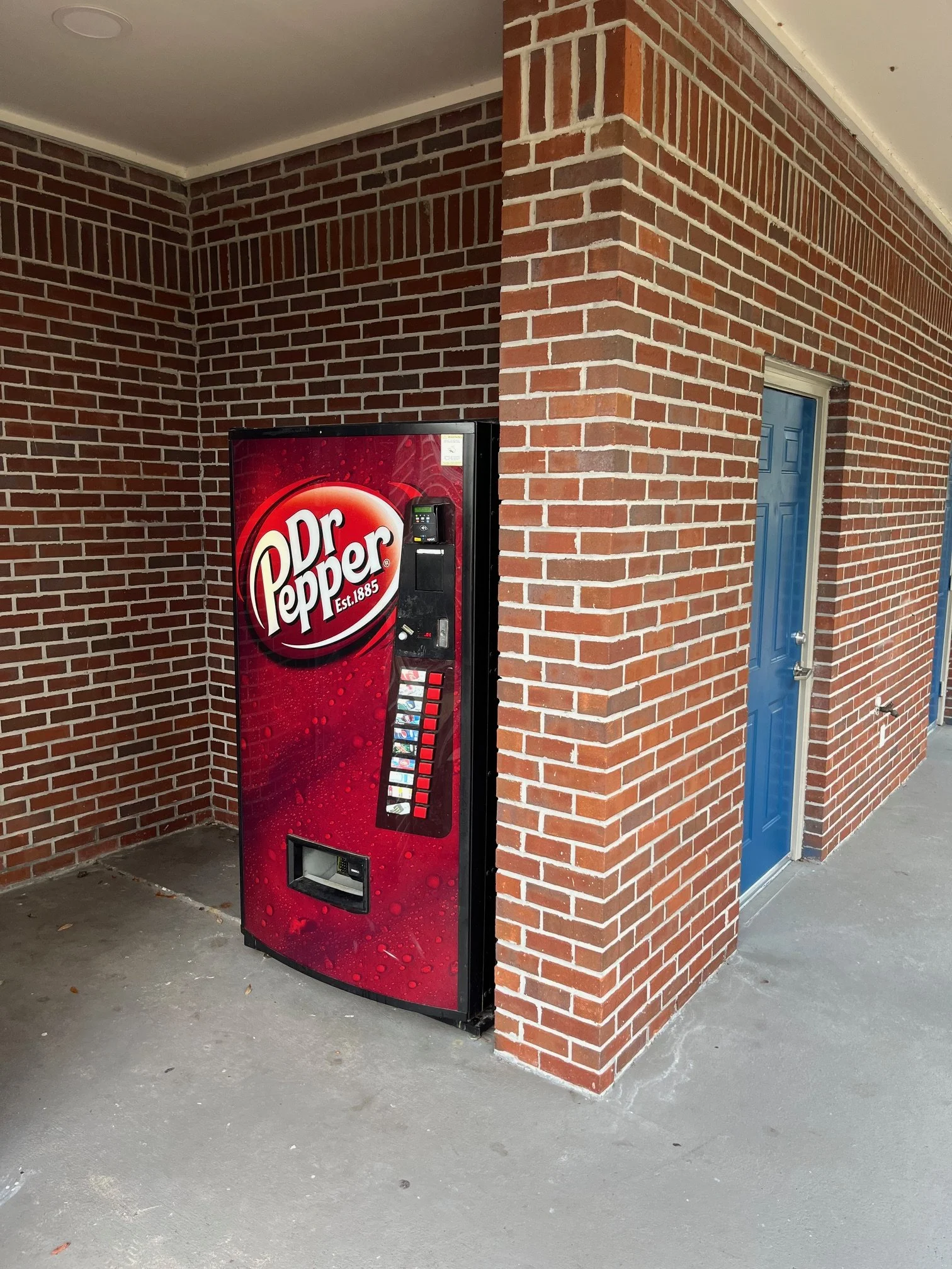 A vending machine for Dr. Pepper soda located outside a brick building with blue doors.