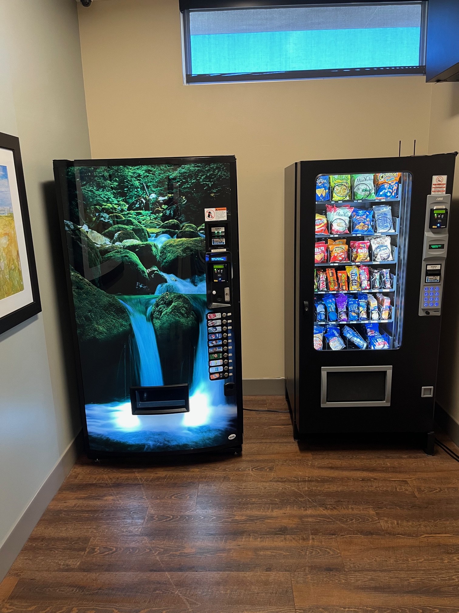 A vending machine with a scenic nature image of a waterfall and rocks, and a snack vending machine filled with chips and snacks, both placed indoors against a beige wall.