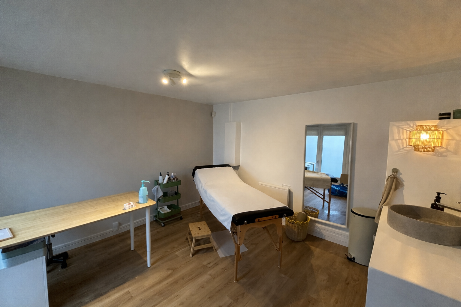 Salle de soins avec table de traitement, lavabo, armoire, et étagère, dans un environnement calme et épuré.