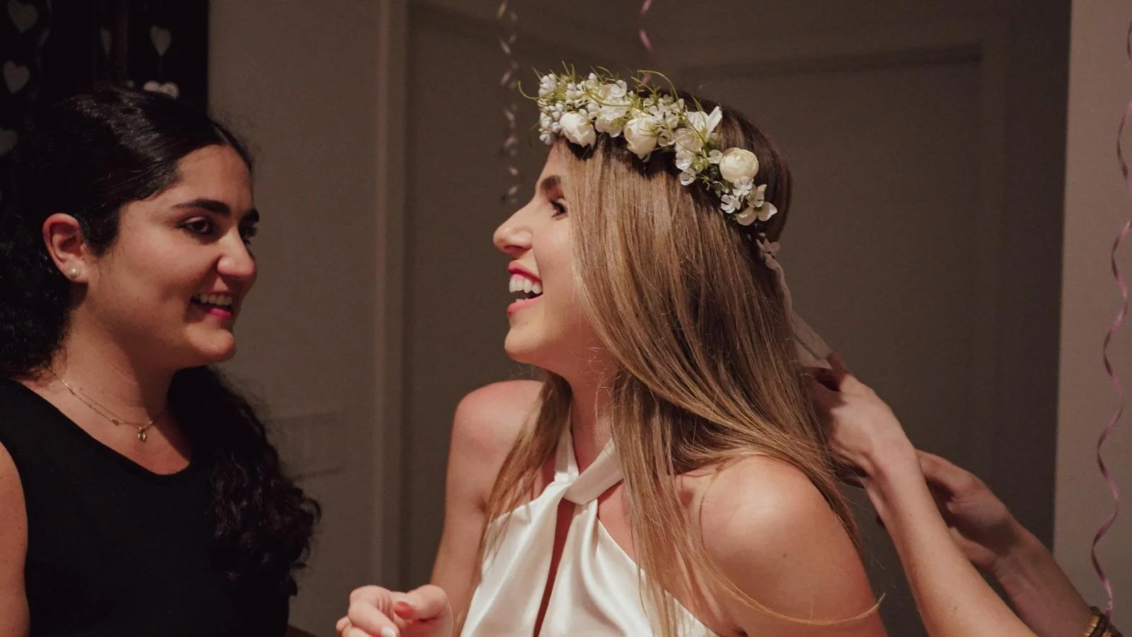 Two women smiling and talking to each other at an indoor event. One woman has dark hair and is wearing a black top, and the other has light brown hair with a floral crown, wearing a white sleeveless top.
