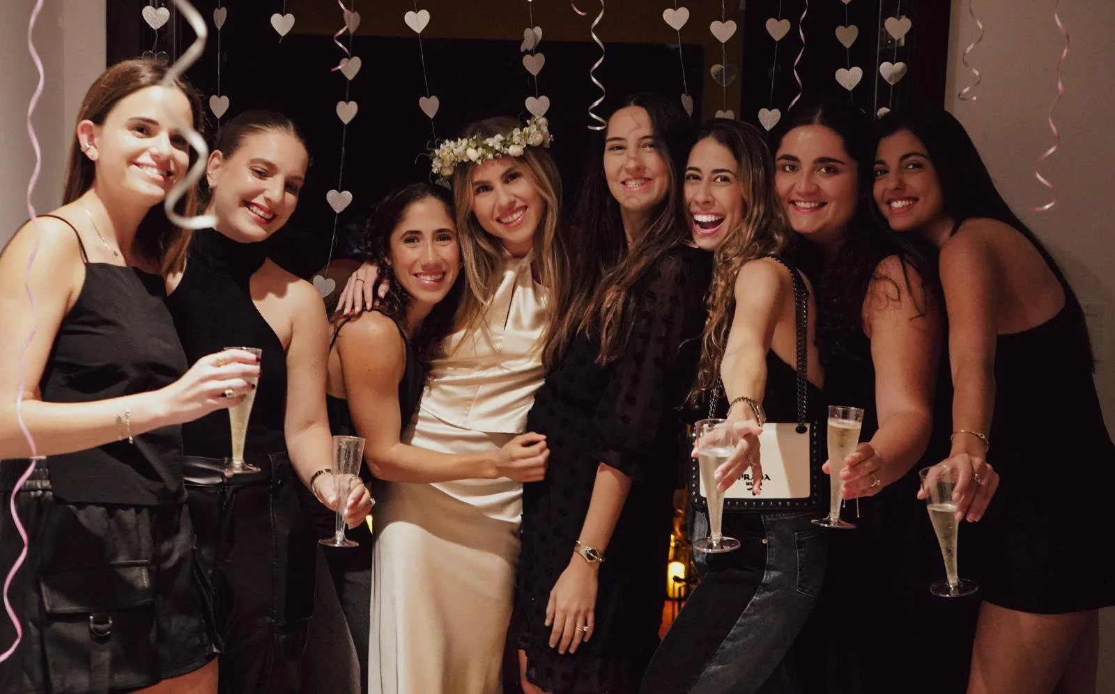 Group of eight women celebrating with drinks, one wearing a flower crown, in a festive setting with hanging heart-shaped decorations.