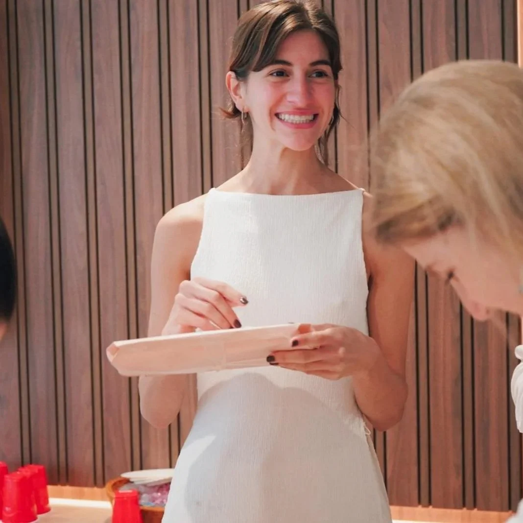 A woman smiling and holding a tray, with another person partially visible in the foreground, in a room with wooden panel walls.