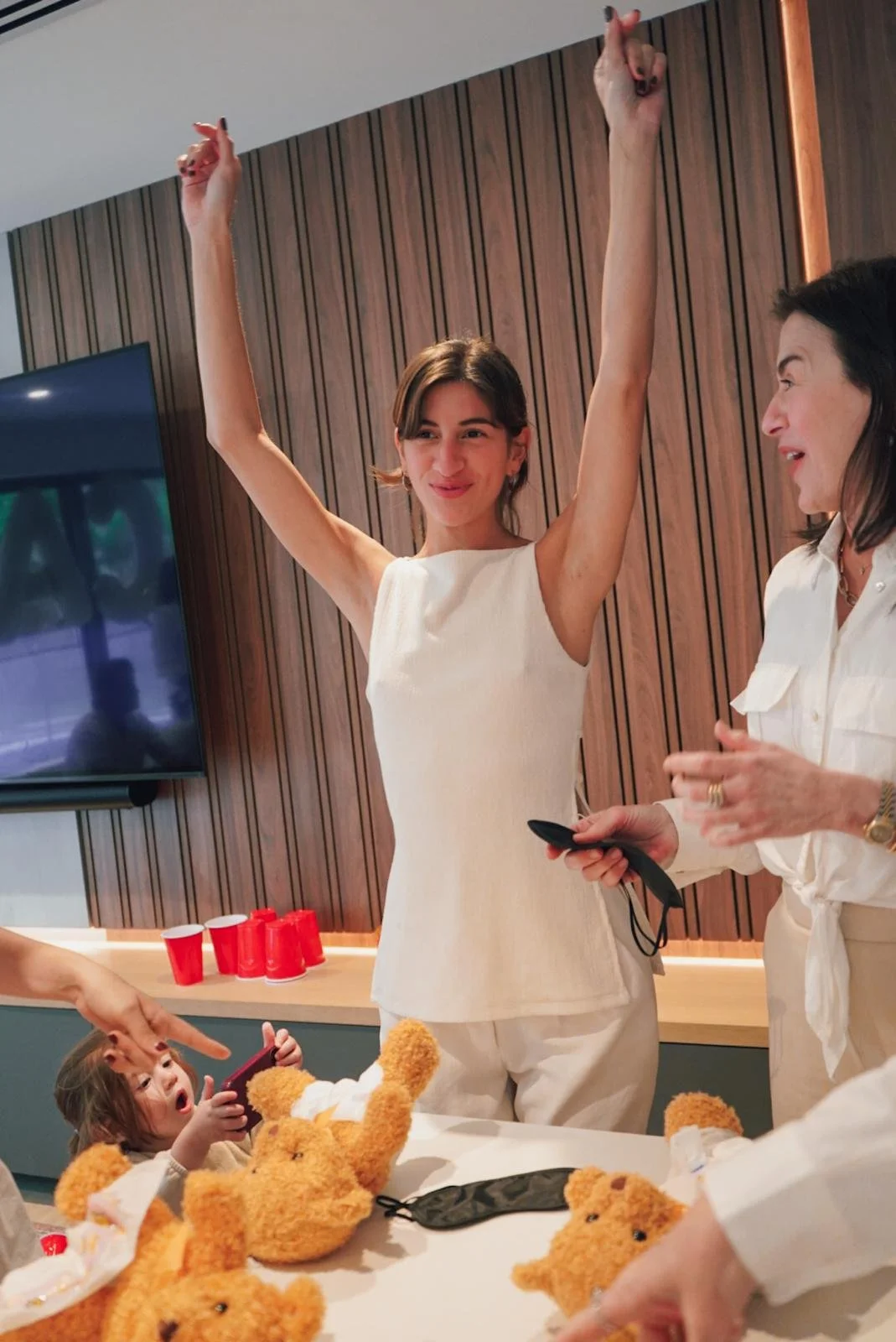 A young woman in a sleeveless white dress has her arms raised in celebration, smiling. She is standing in a room with wooden panel walls, and there are red cups on a sideboard behind her. A small child lying on a table is reaching up towards her whil