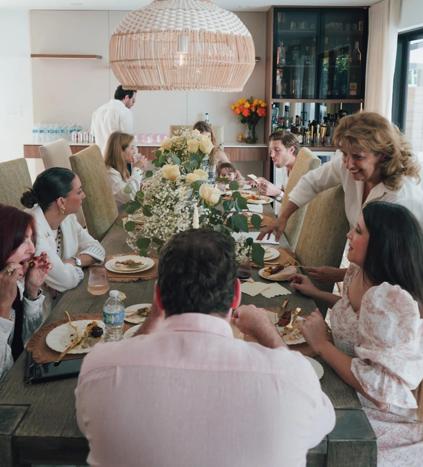 People gathered around a dining table for a meal with floral decorations, in a well-lit room with a bar and large window in the background.