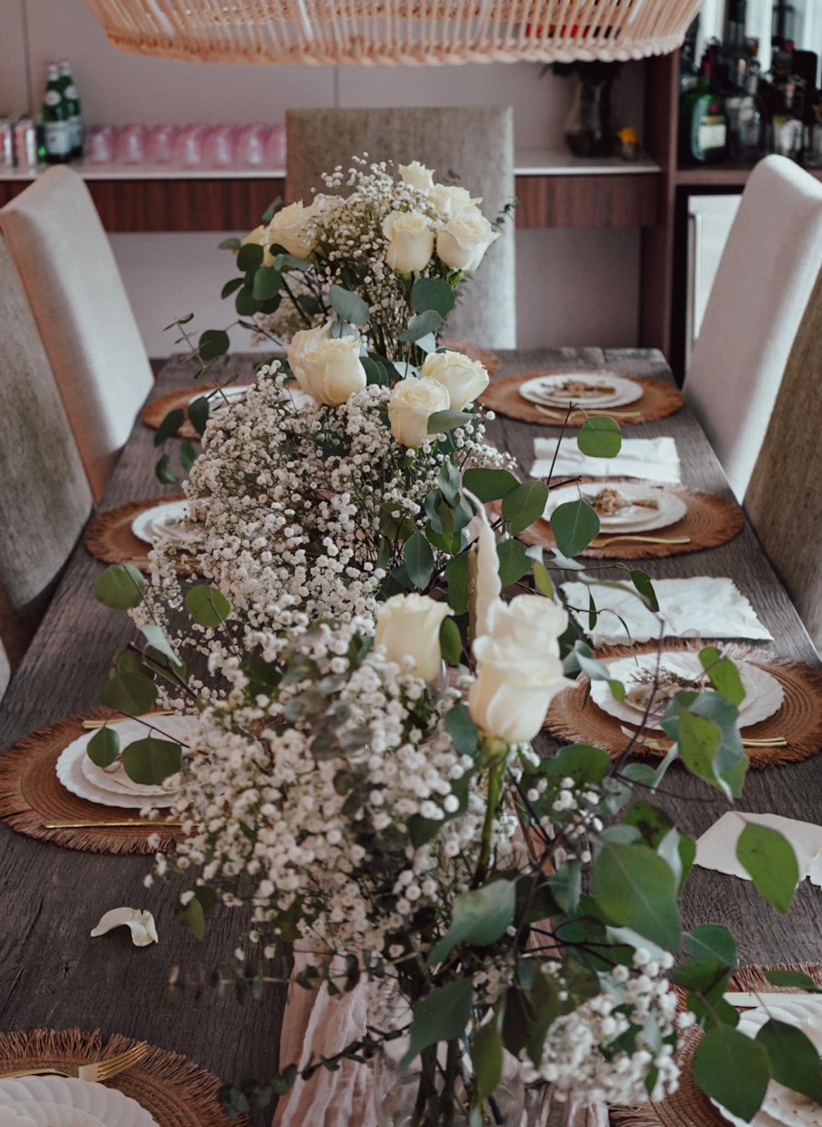 A dining table decorated with white roses, baby's breath, and greenery with place settings and woven placemats.
