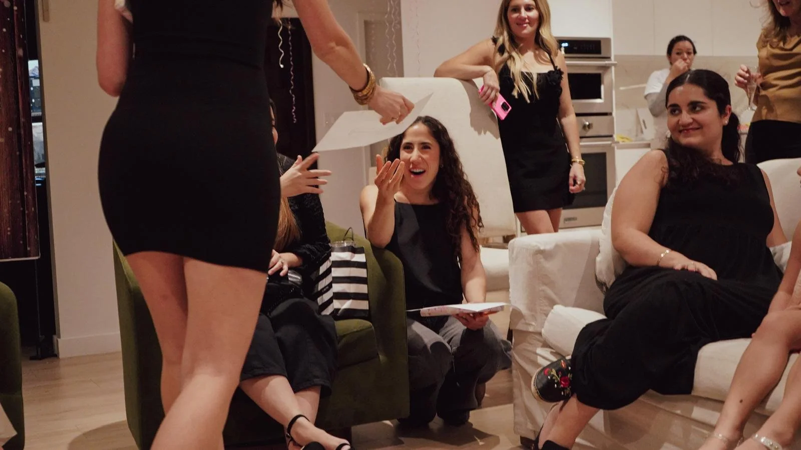Group of women sitting and standing in a living room, enjoying a celebration or party, with one woman reading from a paper.