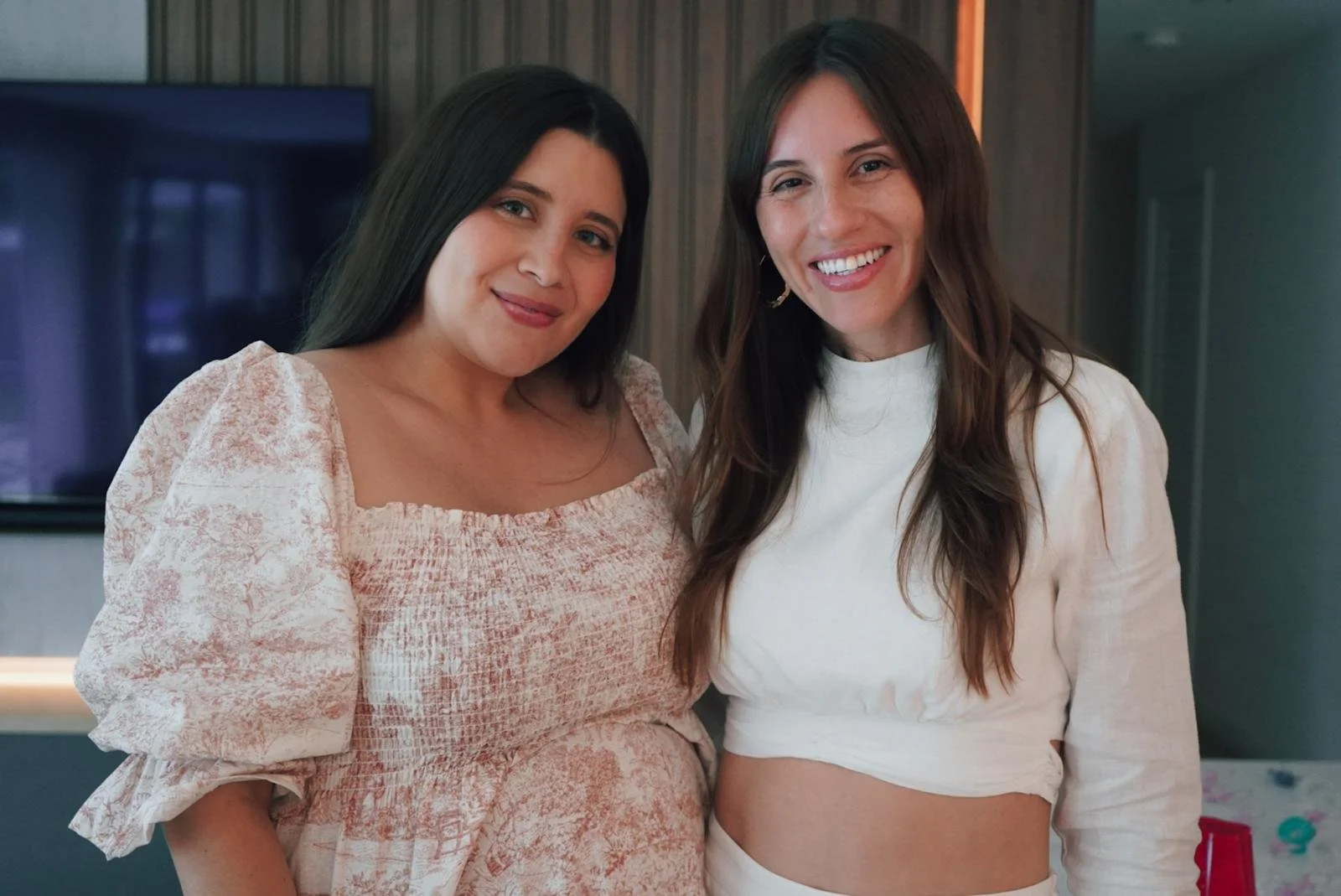 Two women smiling, one with dark hair and a floral dress, the other with light brown hair and a white top, standing indoors.