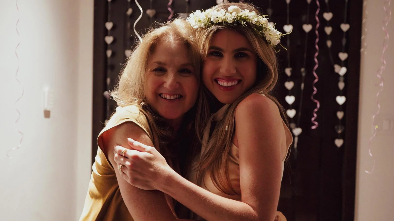Two women hugging and smiling at an indoor celebration, one wearing a flower crown and the background decorated with heart-shaped ornaments and pink streamers.