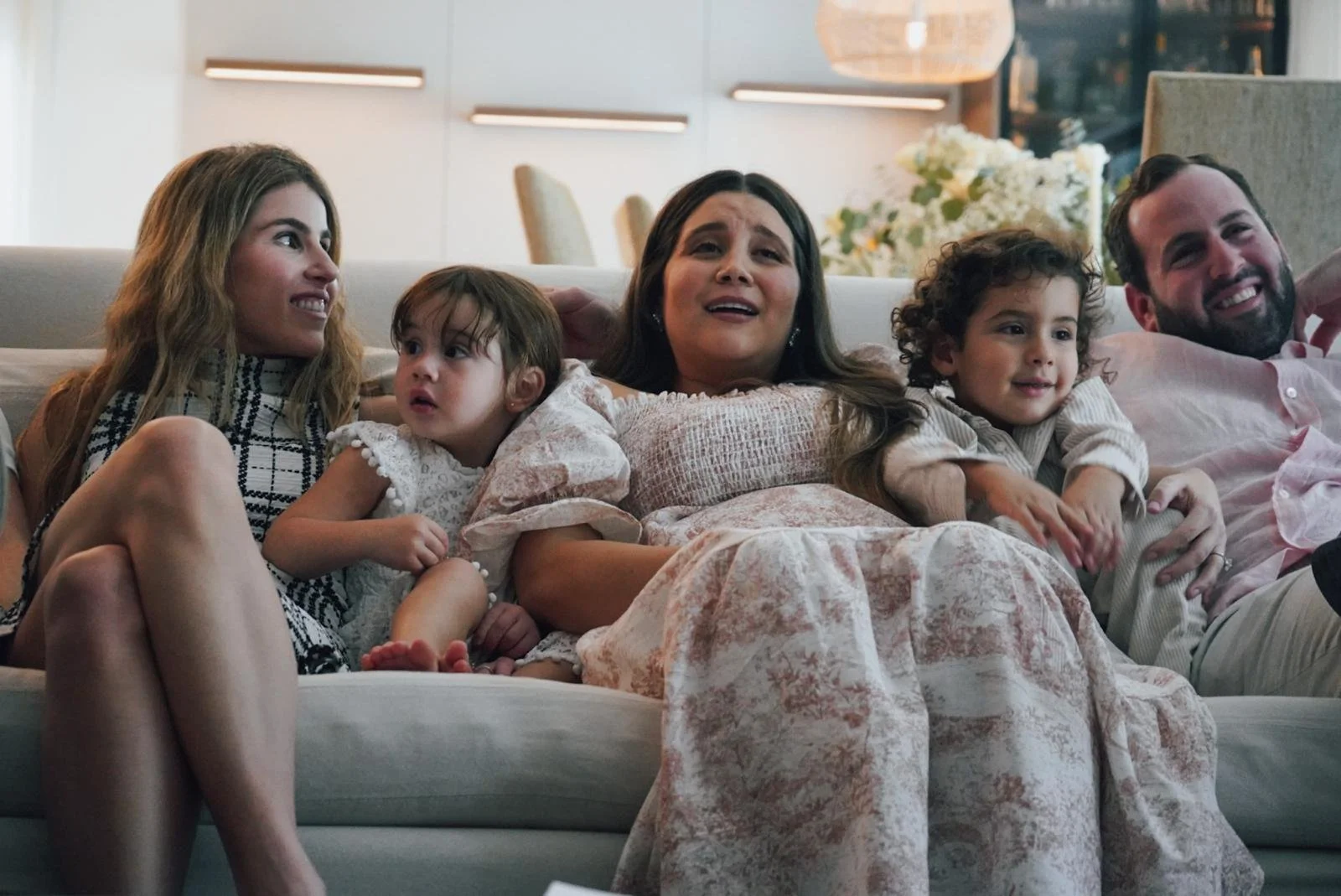 A family of five sitting on a couch, watching television. The group includes a woman and man with three young children, all appearing emotional and engaged.