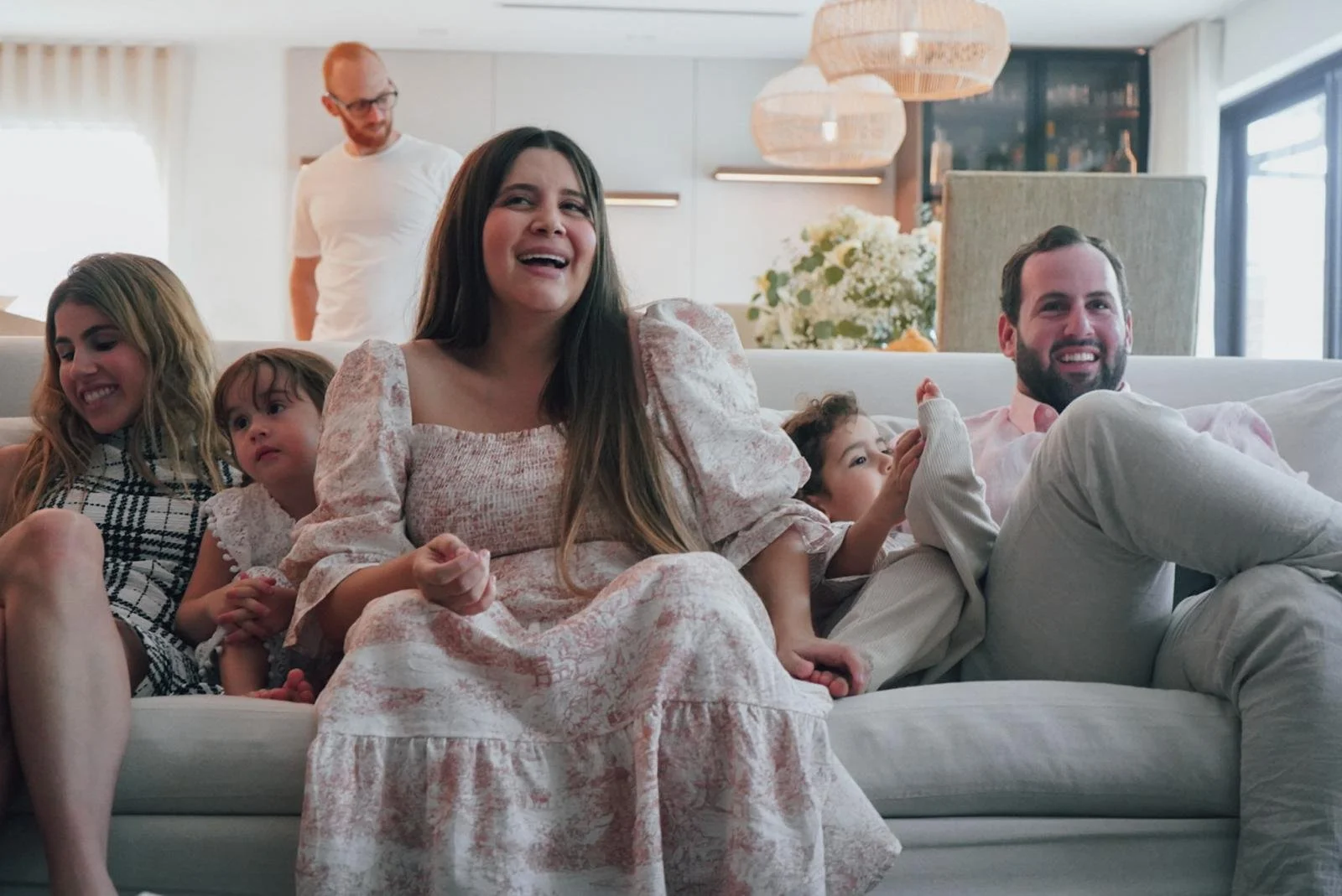 A group of five people, including two children and three adults, sitting on a white couch in a modern living room, laughing and enjoying each other's company.