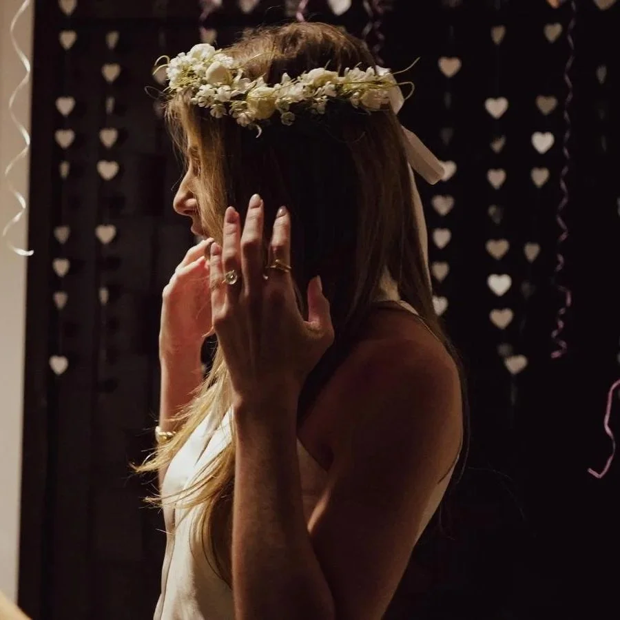 A woman with a flower crown touching her face at a party or celebration, with heart decorations hanging in the background.