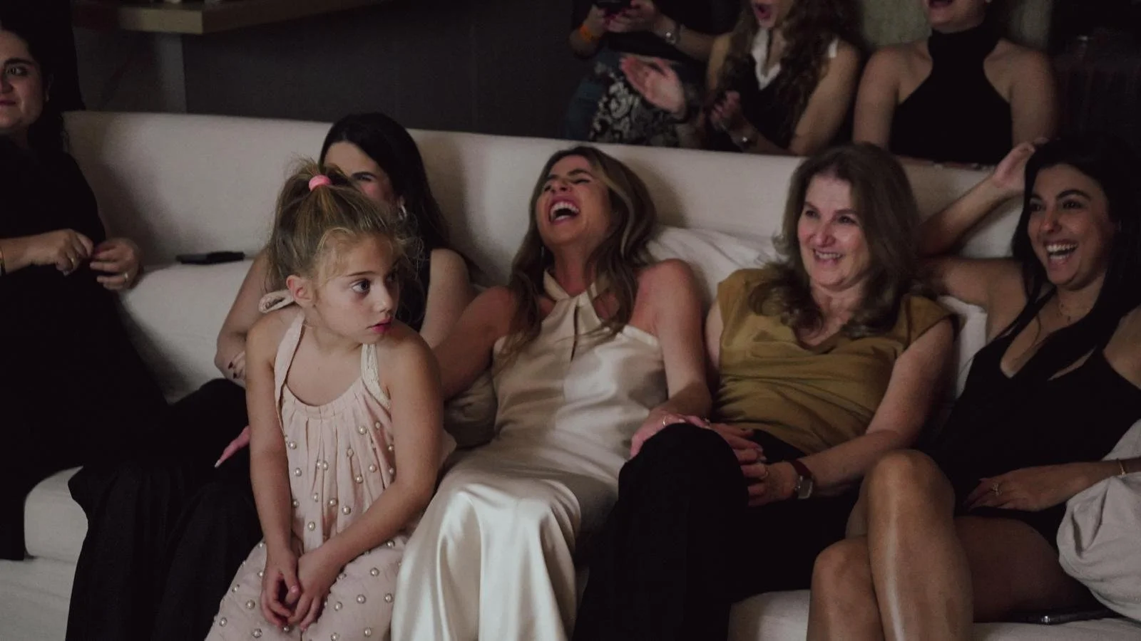 Group of women and a girl sitting on a white couch, laughing and smiling, in a social setting.