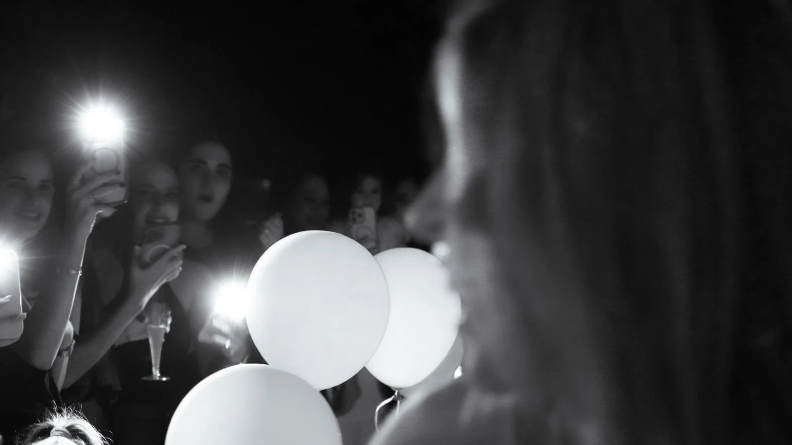 Group of women holding balloons and taking photos at night, some with phone flashlights on.