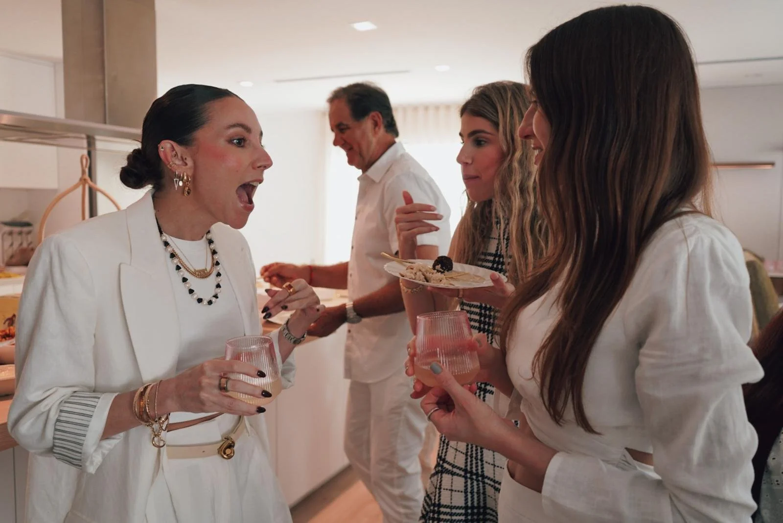 Four women and one man at a social gathering, with three women in the foreground holding drinks and a plate of food, engaging in conversation. The woman on the left, dressed in white, is speaking animatedly.