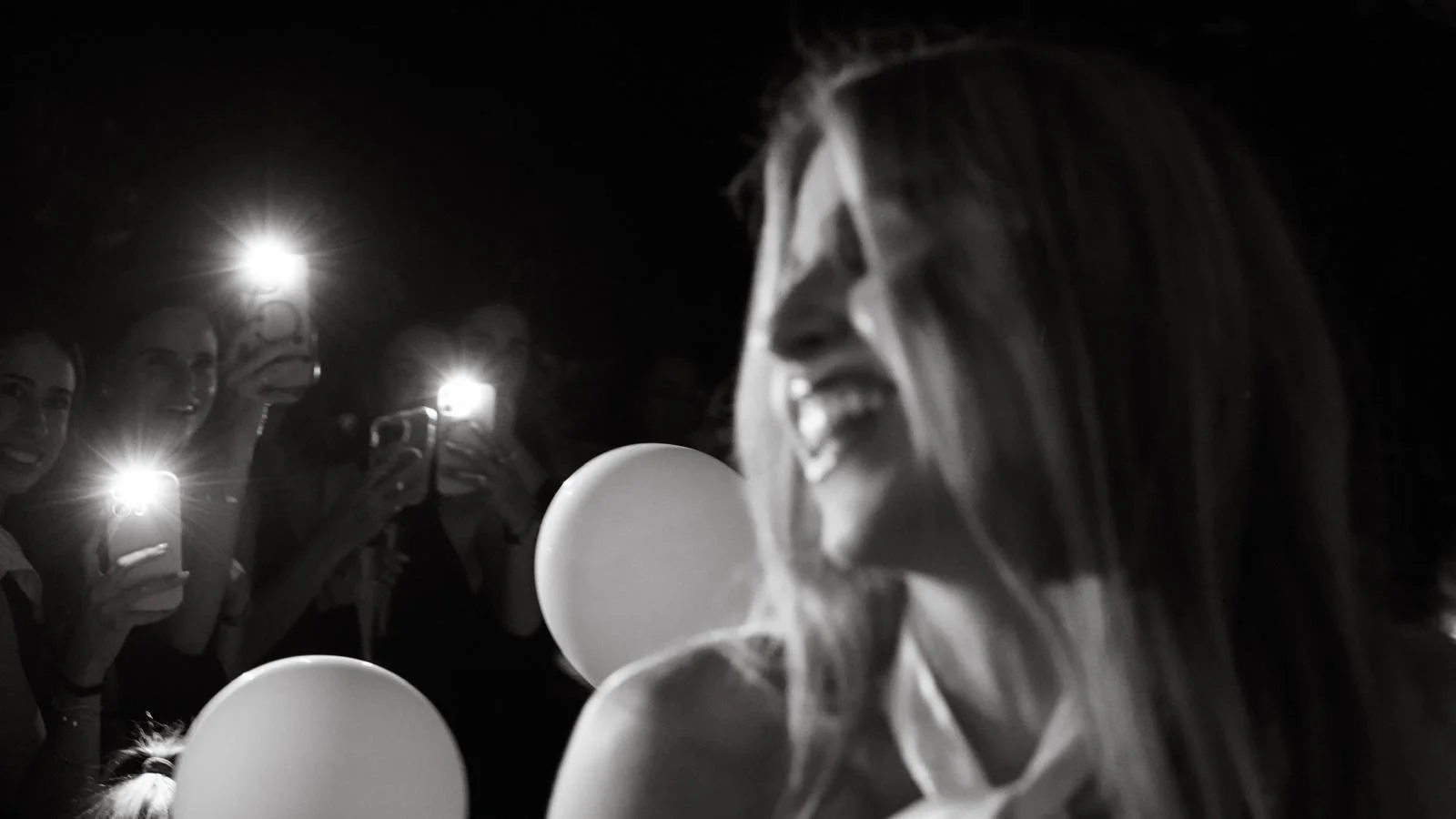 A woman laughing with balloons in the foreground and several people taking pictures with their smartphones in the background at night.