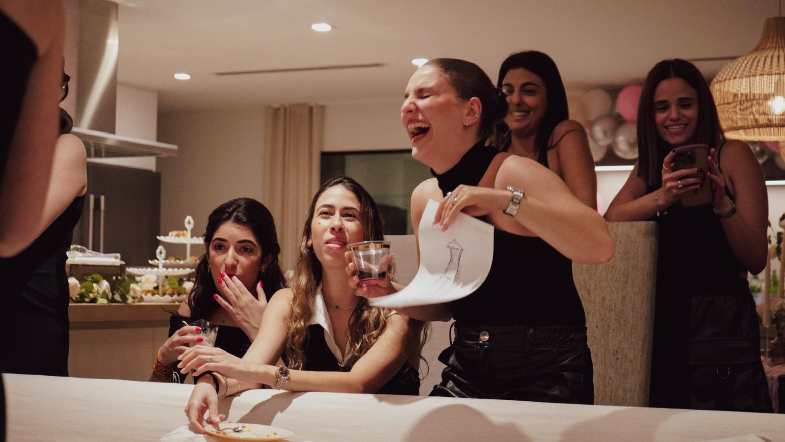 A group of women gathered around a table at a celebration, with one woman laughing and holding a drink and a paper, while others look on and smile, in a warmly lit room.