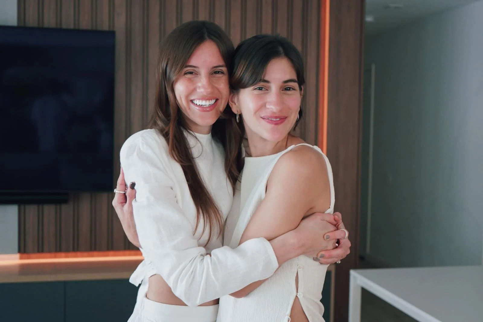 Two women hugging and smiling at the camera in an indoor setting with a wooden wall and a large television behind them.