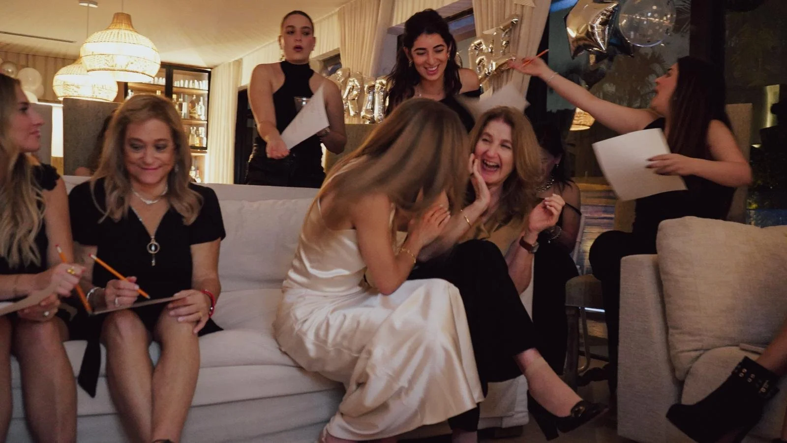 Group of women celebrating at a party, with balloons, gifts, and decorations in the background.