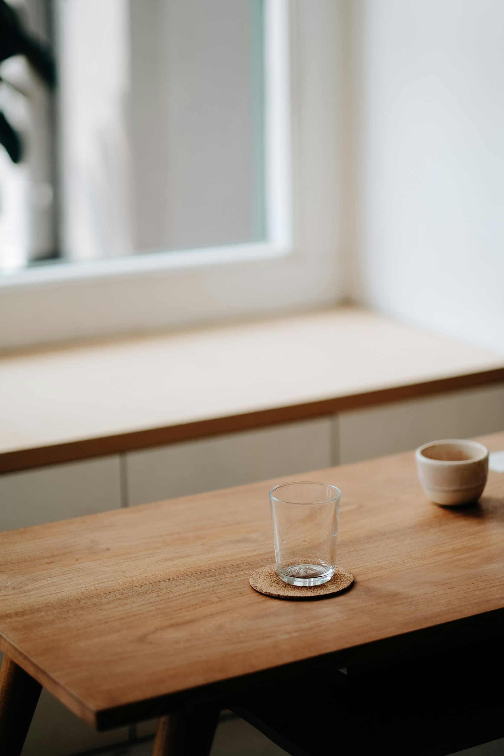 Empty glass on a cork coaster and a bowl on a wooden table near a window with a white frame.