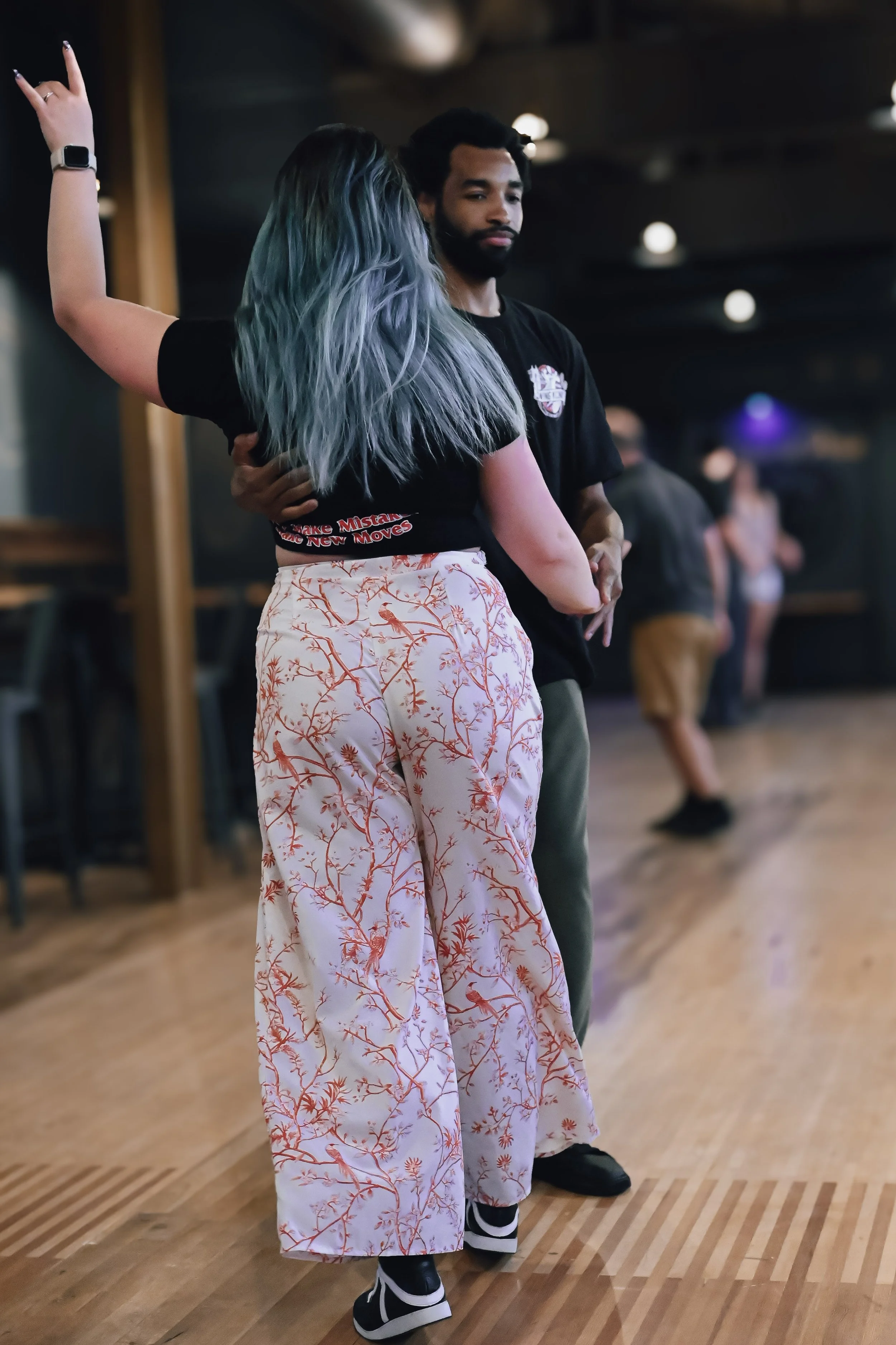 A woman with long blue and purple hair dancing with a man in a black shirt in a dimly lit indoor space with a wooden dance floor.