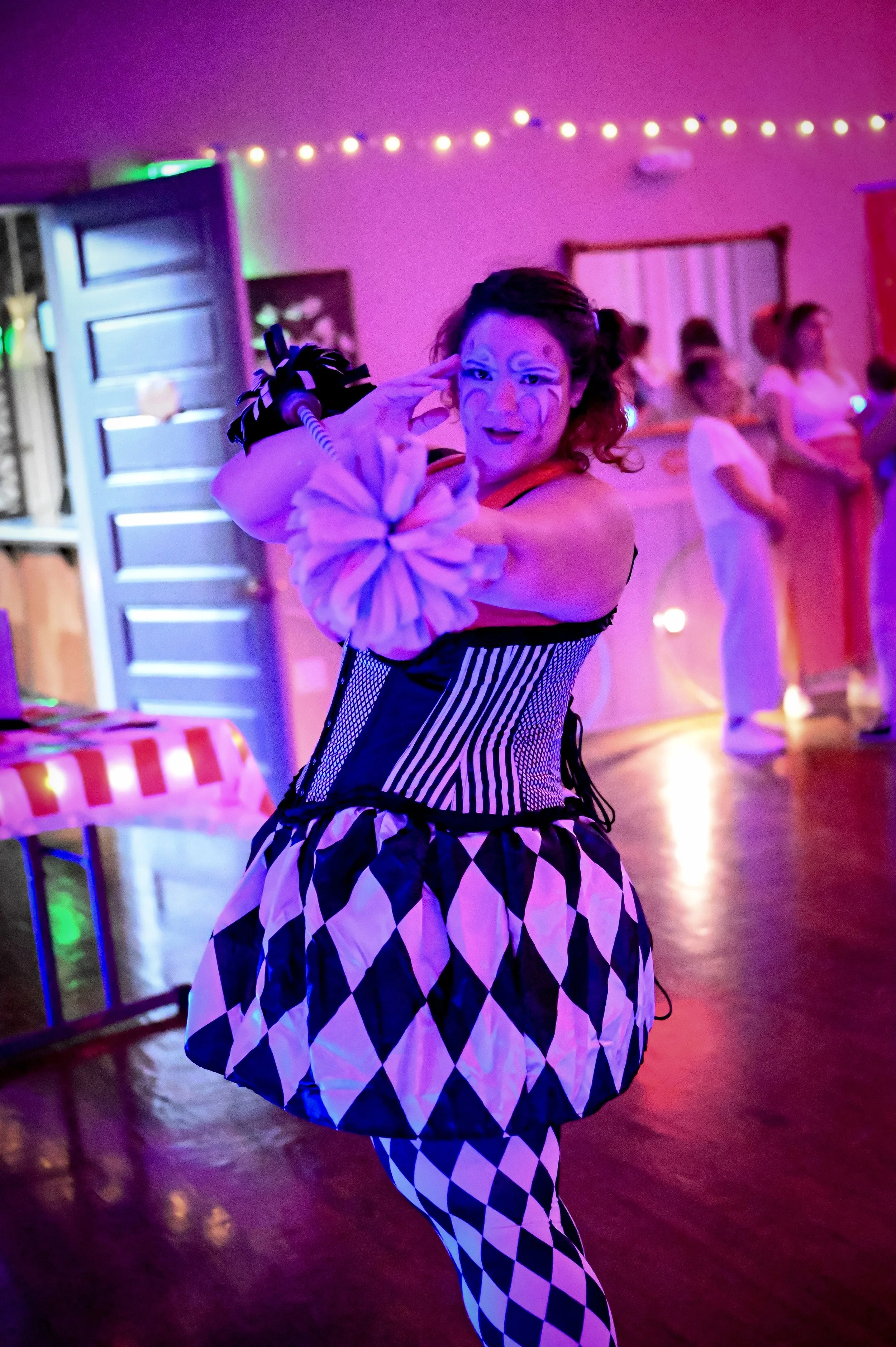 A performer dressed as a clown with face paint, striped corset, and harlequin-patterned skirt and leggings, striking a playful pose at an indoor party with pink and purple lighting.