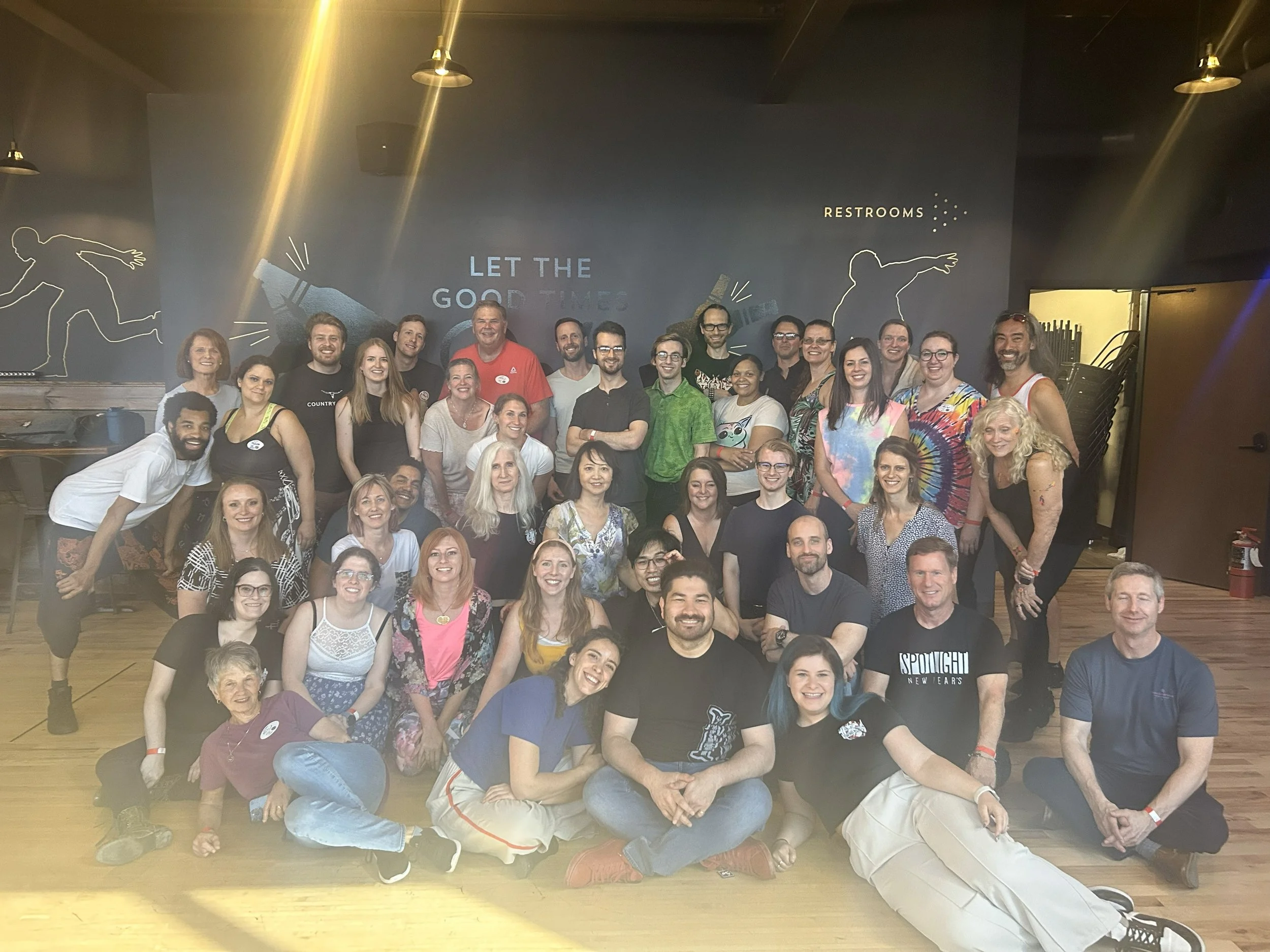 A large group of smiling people gathered together indoors, posing for a photo in front of a wall with the words "LET THE GOOD TIMES" and "RESTROOMS." The group is diverse in age and ethnicity, and they appear happy and friendly.