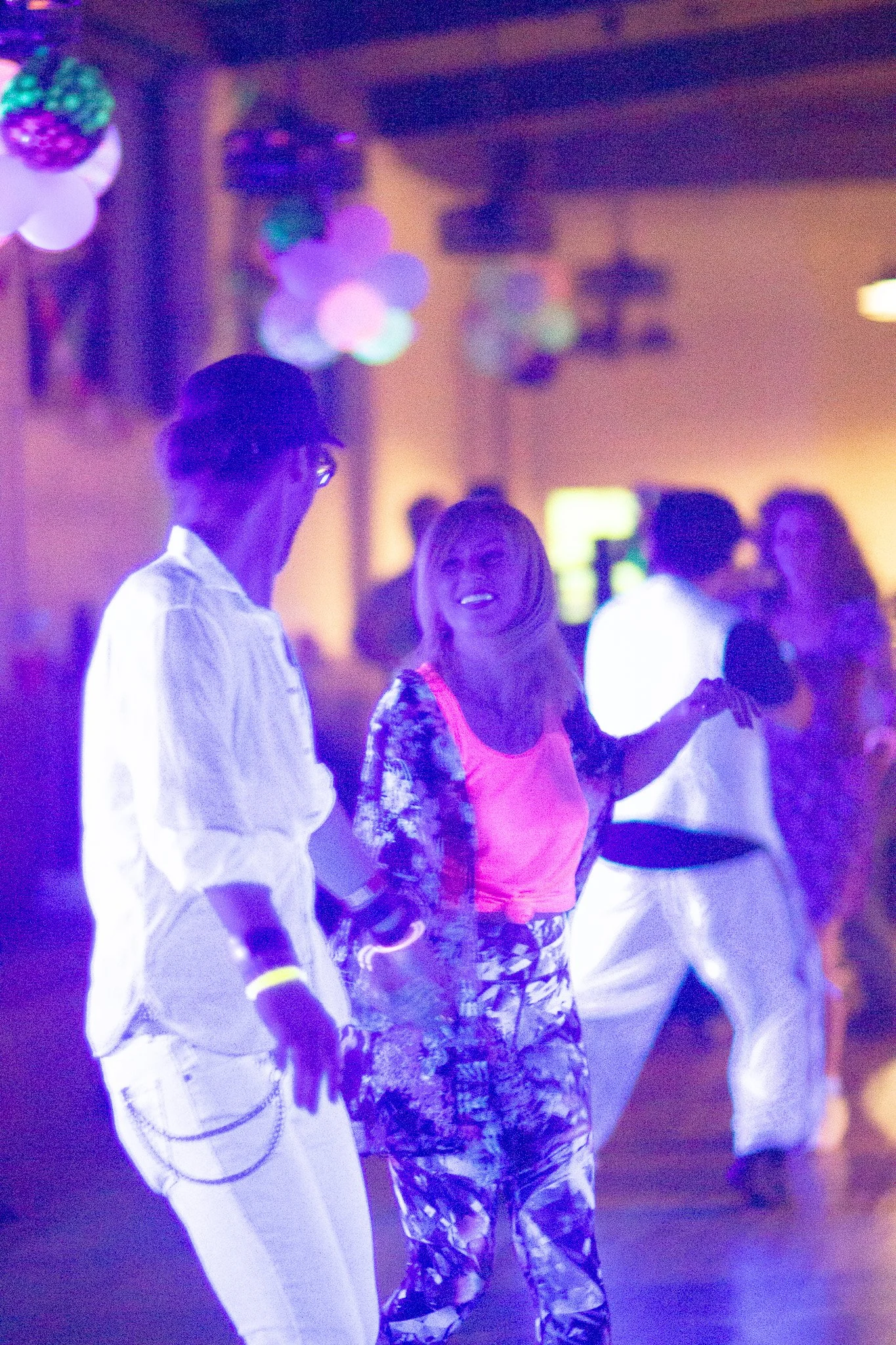 People dancing at a party with colorful balloons and decorations, illuminated by purple and pink lights.