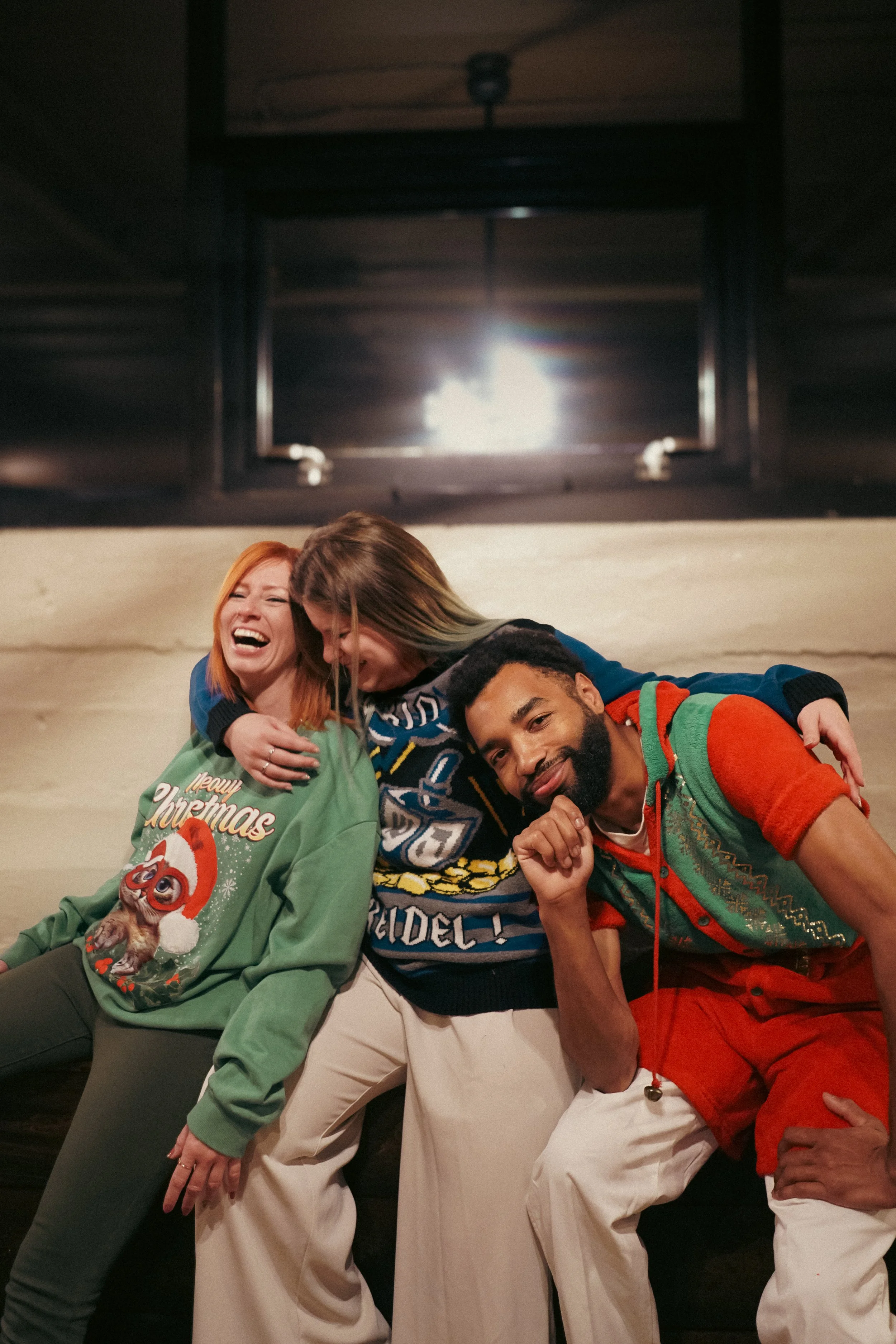 Three people sitting close together, laughing and smiling, wearing Christmas sweaters, in a cozy indoor setting.