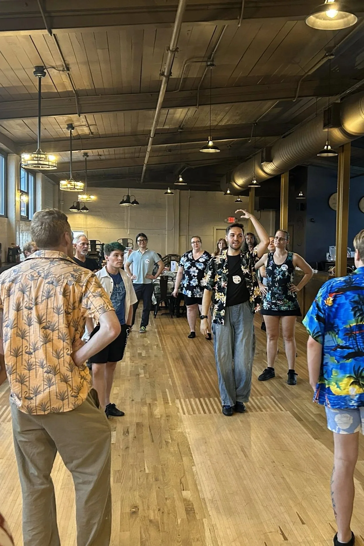 People wearing tropical shirts participating in a dance class or social event indoors, with wooden floors and a wood-paneled ceiling.
