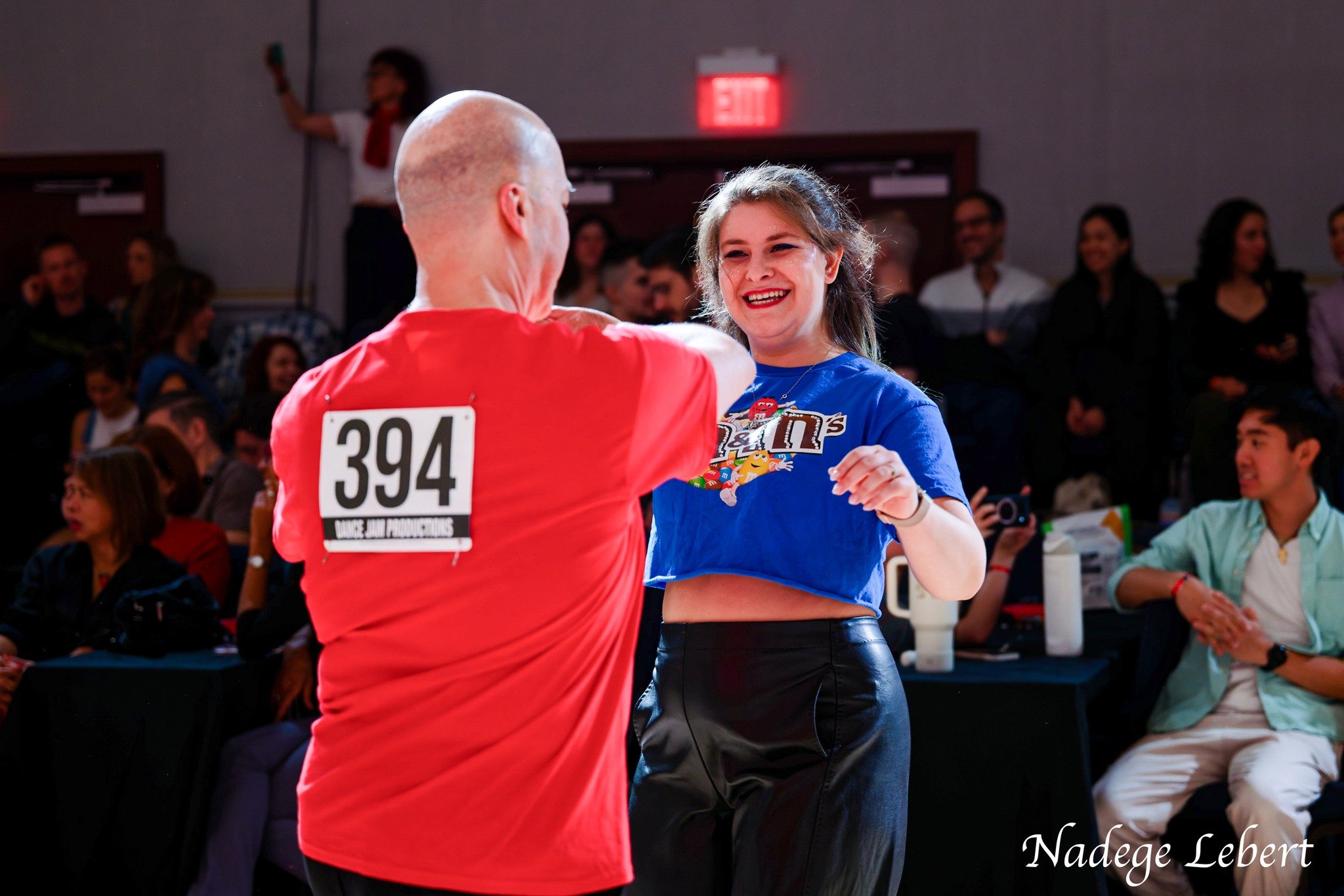 A man and a woman dancing at a lively indoor event, with the man wearing a red shirt and the woman in a blue crop top and black pants, surrounded by seated and standing spectators.