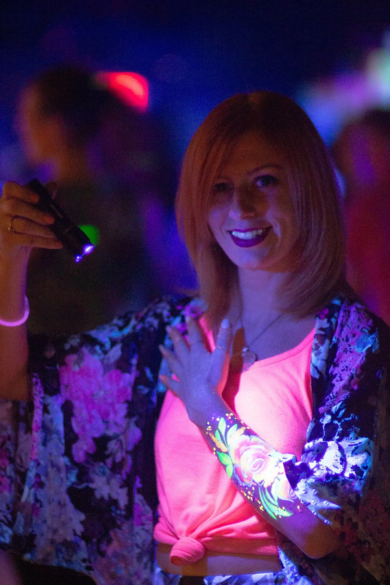 A woman in a neon-lit environment holding a small flashlight, smiling, with colorful glow-in-the-dark body paint on her arm.