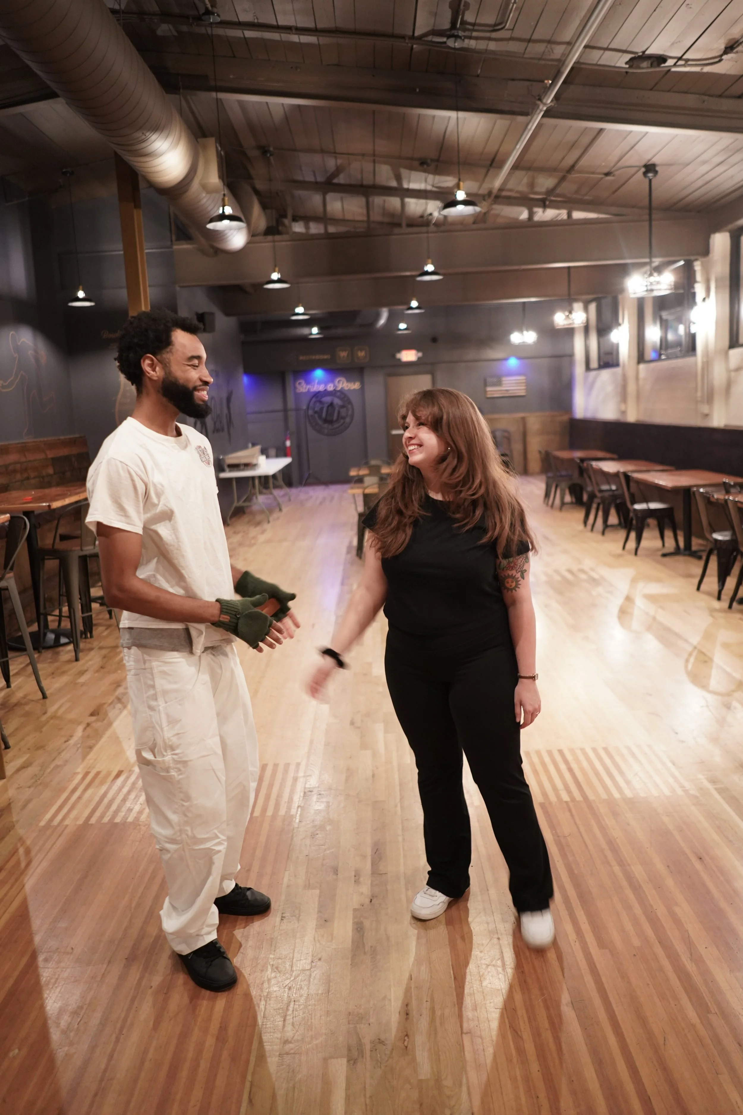 A man and woman are smiling and talking in a spacious, dimly lit restaurant or bar with wooden flooring and chairs along the walls.
