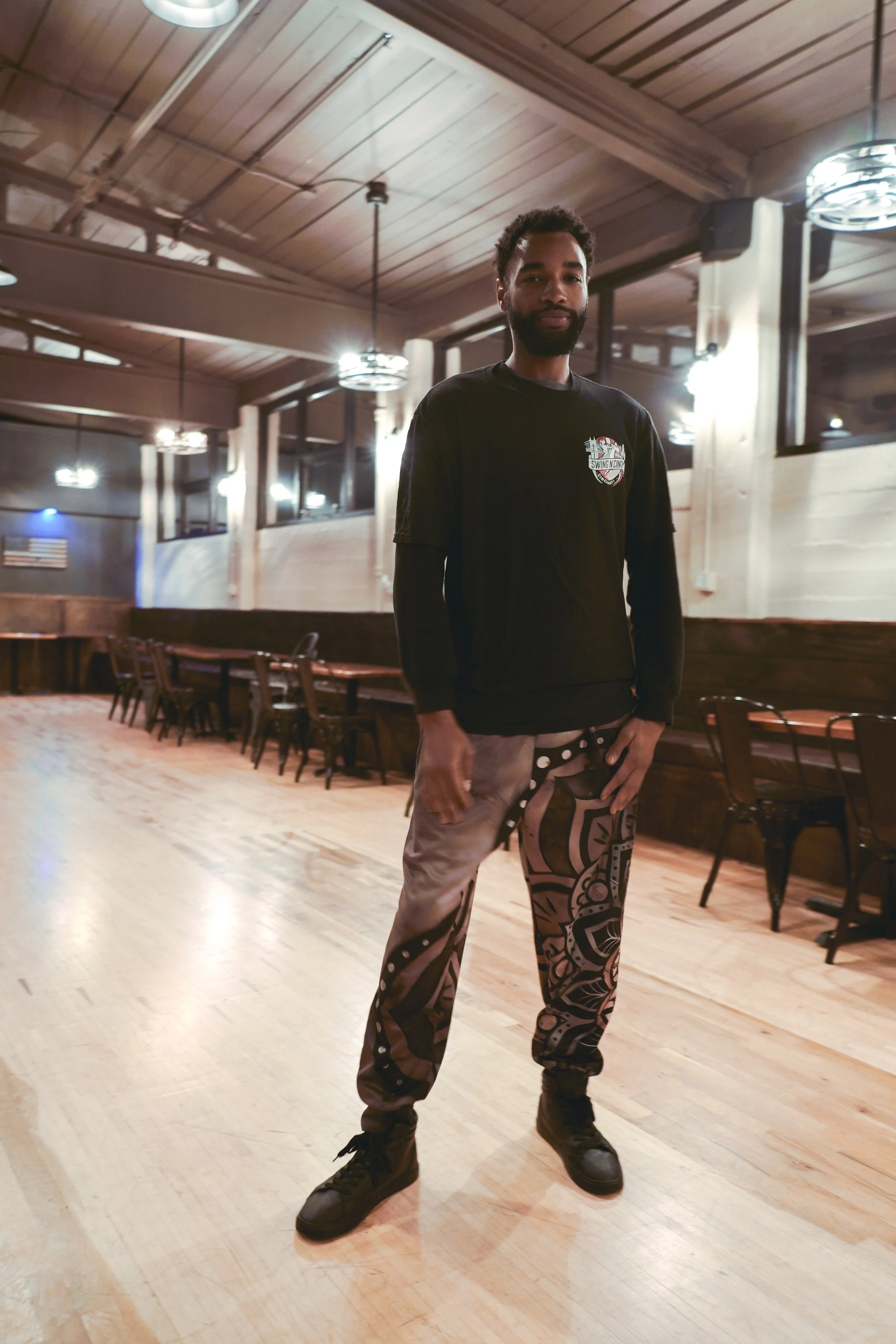 A man with a beard and dark hair standing in a dimly lit wooden room with tables and chairs, wearing a black long-sleeve shirt and patterned pants.