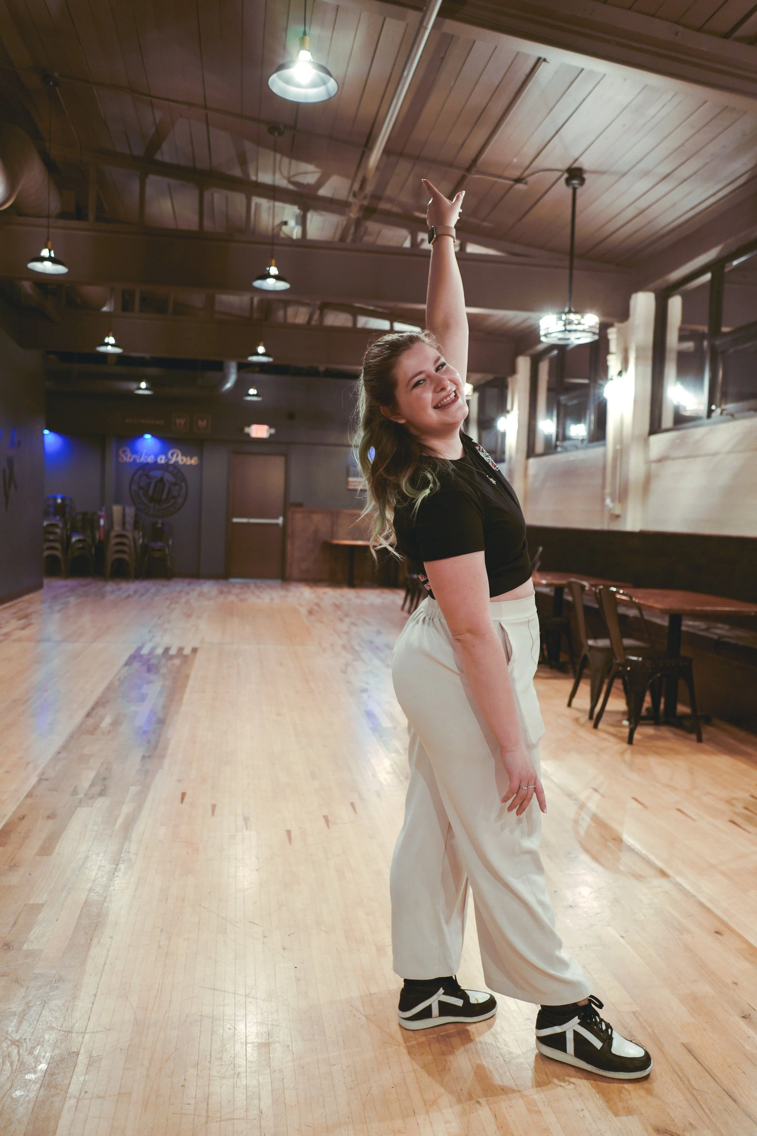 A young woman dancing in a wooden-floored dance studio, smiling, with one arm raised and the other resting on her leg.
