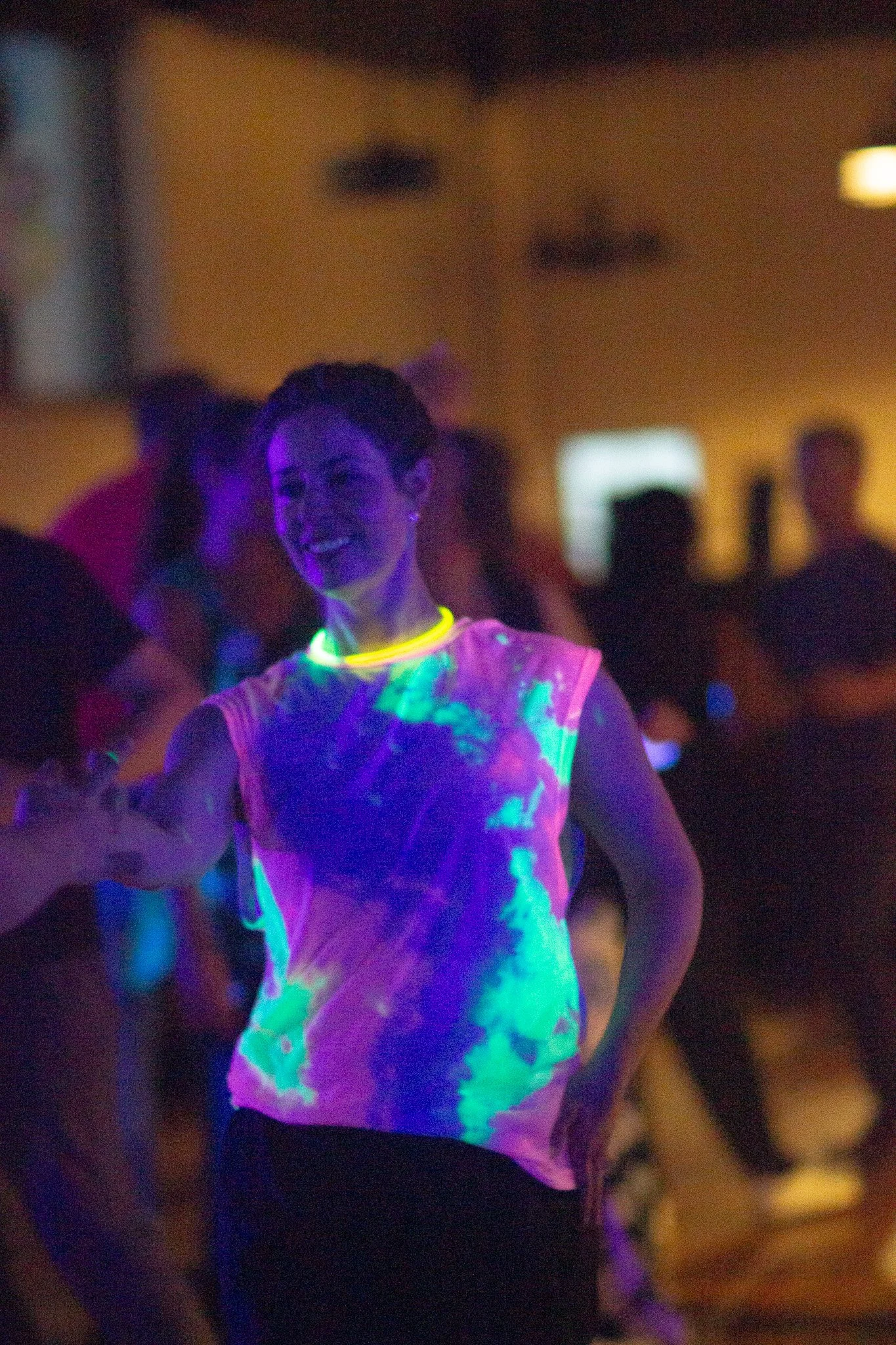 A woman smiling and dancing at a party with black lights, wearing a tie-dye shirt that glows under the lights.